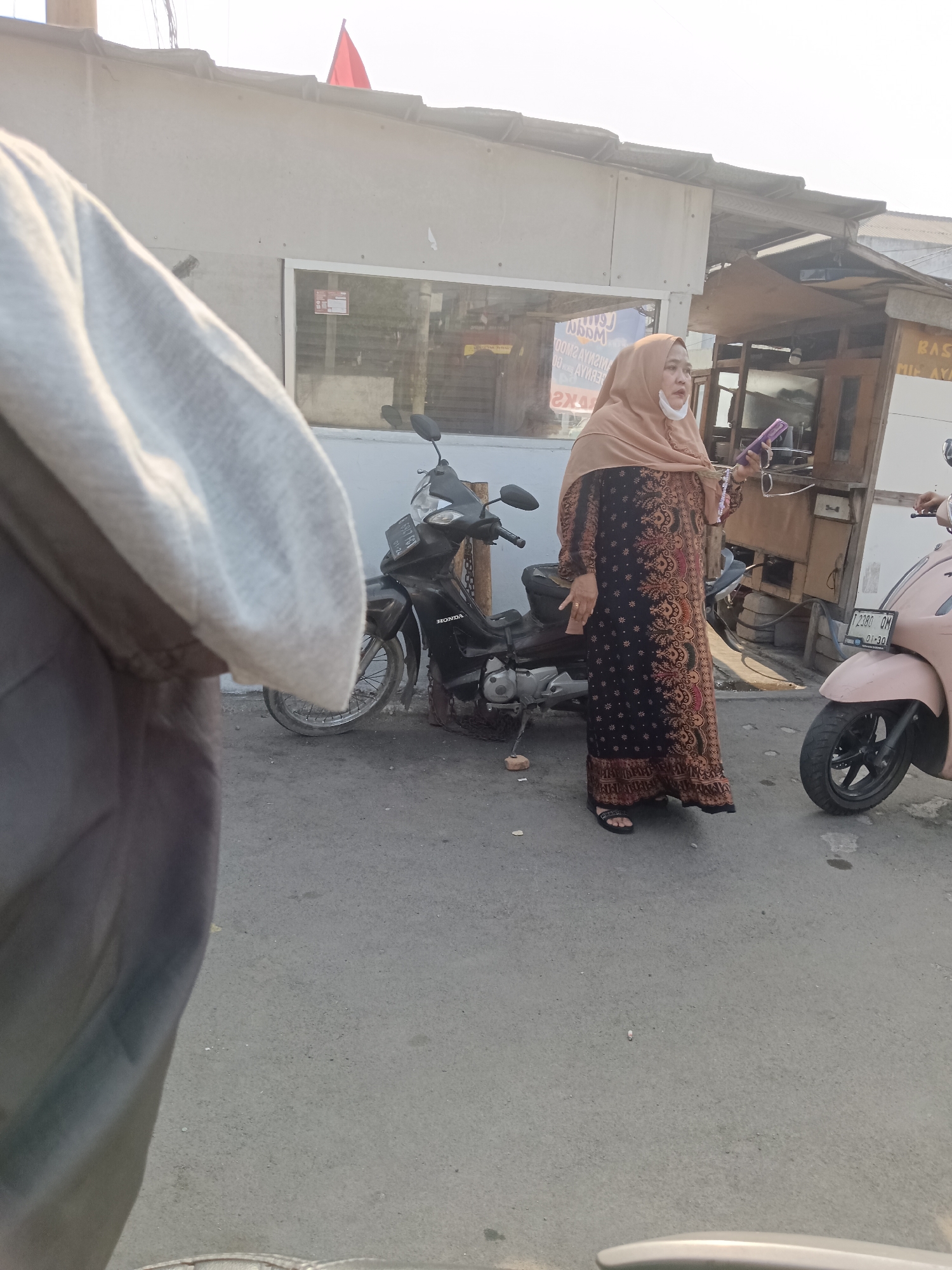 The image captures an outdoor scene in Kedungwaringin, Indonesia, featuring a woman standing on a paved road. She is wearing a light brown hijab and a long, dark patterned dress with gold and red accents, along with black sandals. The woman is holding a purple object in her right hand and has a white mask loosely covering her chin. Behind her, a black motorcycle is parked, and to her right, a light pink scooter is also visible. The background shows a building with a light-colored facade and a corrugated metal roof. The lighting suggests it is daytime, possibly late afternoon, with sunlight casting shadows. The overall environment appears to be an urban or semi-urban area, consistent with a commercial or residential street in Indonesia.