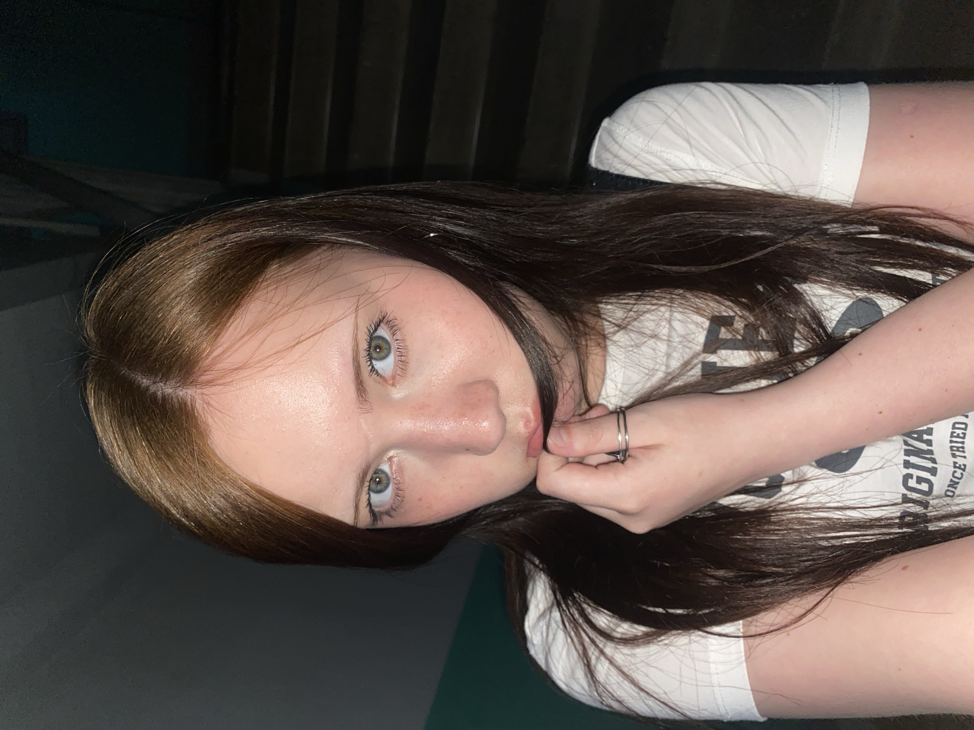 A young woman is posing for a photo. She has light skin, brown hair, and green eyes. She is wearing a white t-shirt. Her right hand is near her face, and she is wearing two rings on her fingers. The background appears to be a stairway. The location is Unknown, Unknown.