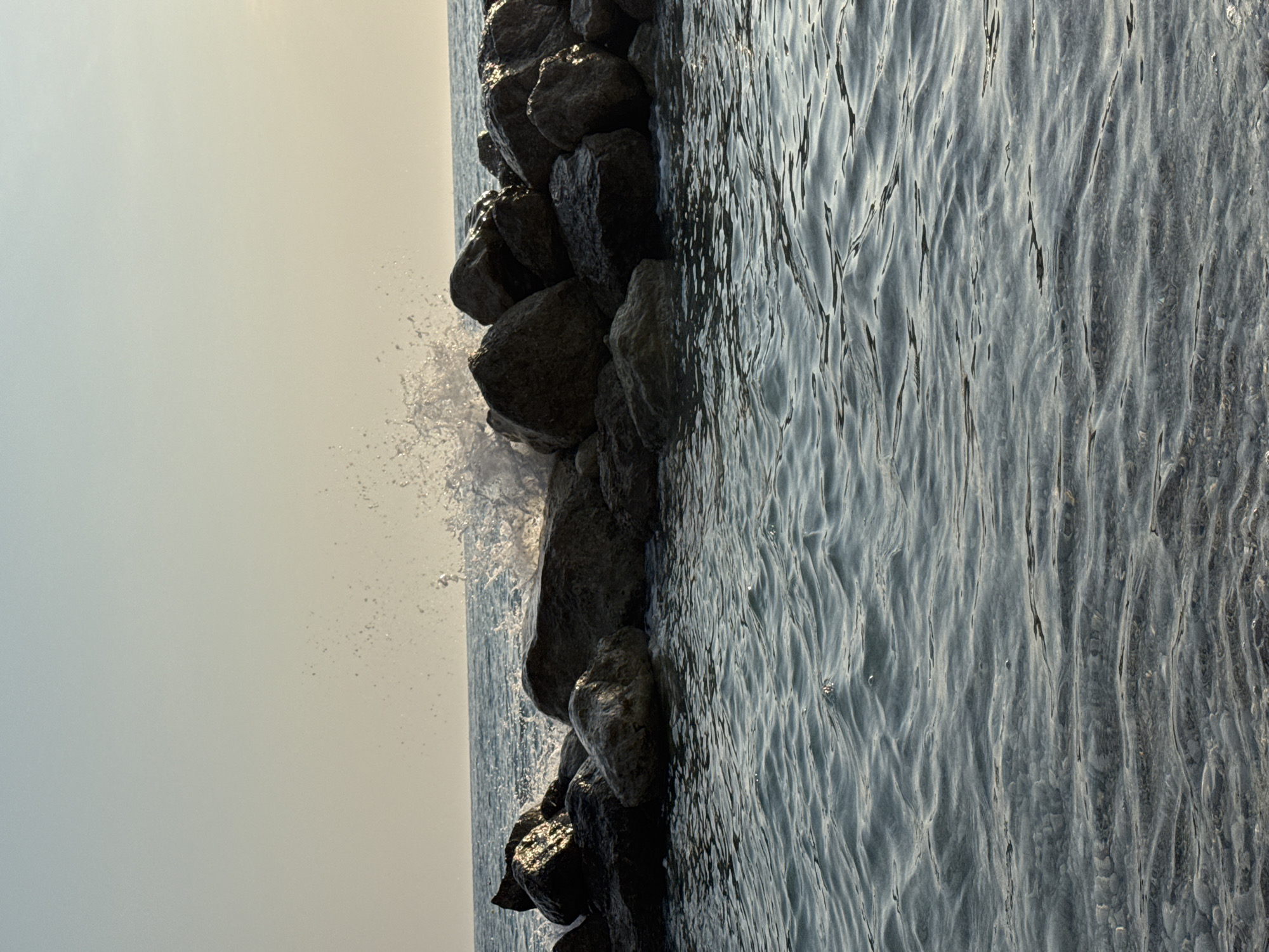 The image depicts a serene seascape with a rock formation jutting out into the water. Waves are seen crashing against the rocks, creating a dynamic splash of white water. The sky above is a soft gradient of pale blue and beige, suggesting either early morning or late afternoon light. The water in the foreground is calm, with gentle ripples reflecting the muted tones of the sky. The scene is likely located along the coast of Fethiye, Turkey, a region known for its beautiful Mediterranean beaches and rocky shores. There are no people visible in the image.