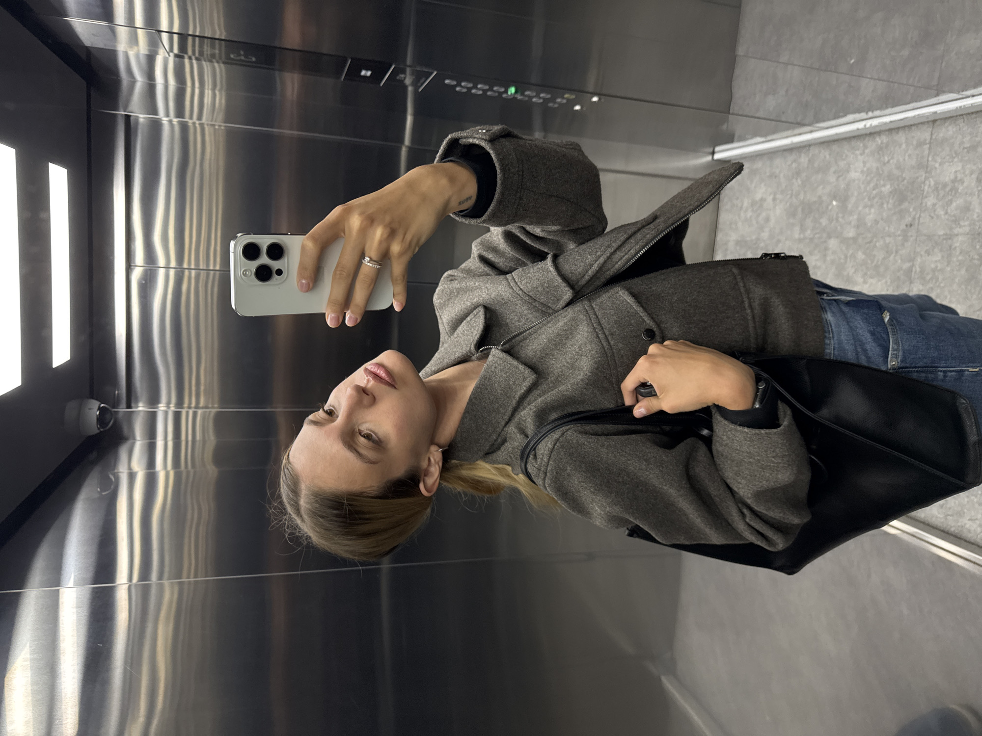 A young woman is taking a selfie in an elevator. She is holding a white smartphone in her left hand. She is wearing a gray coat, a black long-sleeved shirt, and blue jeans. She has a black bag on her shoulder. The elevator has stainless steel walls and a control panel with buttons and lights. The floor is light gray tile. The location is in an unknown city.