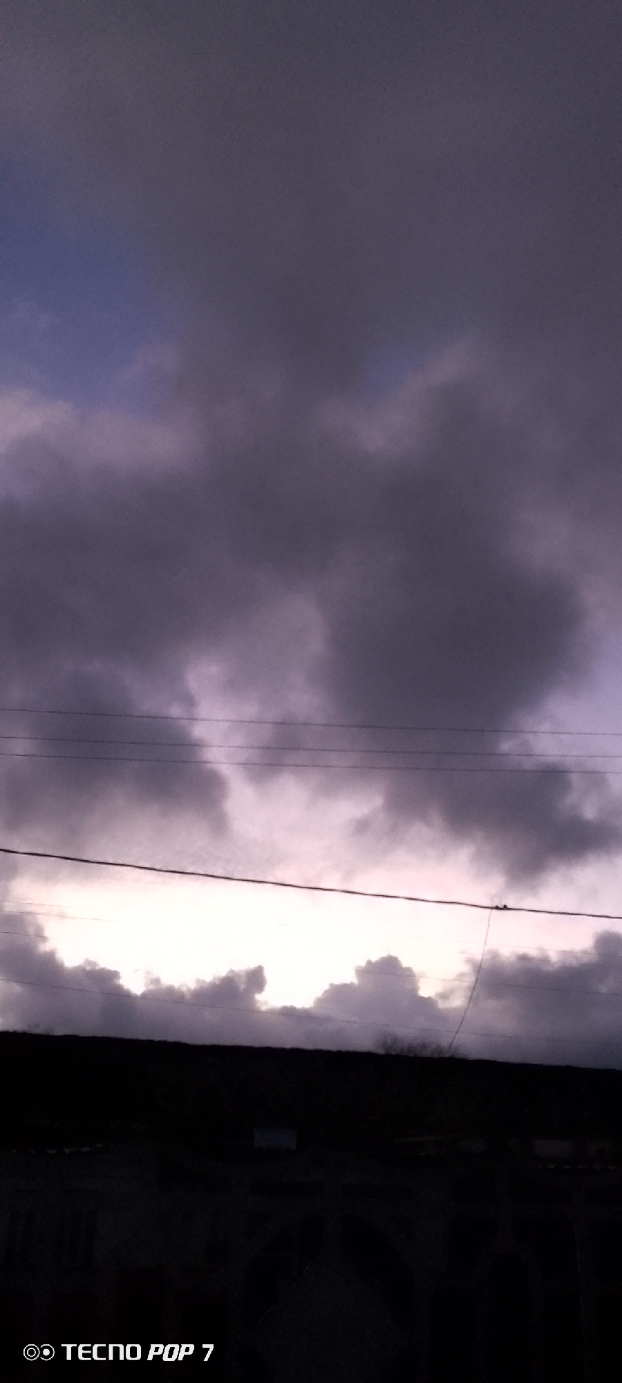The image presents a dramatic twilight sky over Altagracia, Venezuela. The scene is largely dominated by a vast expanse of heavy, dark grey and purplish clouds that fill the upper and central portions of the frame, suggesting overcast or stormy weather conditions. A striking bright, horizontal band of light cuts across the lower middle of the sky, likely indicating the sun's position during either dawn or dusk, or sunlight breaking through a dense cloud layer.
Several thin, dark power lines or telephone wires stretch horizontally across the mid-ground, silhouetted against the bright band of light and the surrounding darker clouds. Below these wires and the bright horizon, a dark, indistinct silhouette of what appears to be buildings or a distant landscape is visible, further emphasizing the low light and atmospheric conditions. No people or specific activities are discernible in the photograph. The overall color palette is muted, consisting primarily of deep greys, purples, and faint blues in the sky.
In the bottom left corner of the image, the text "TECNO POP 7" is clearly visible, indicating the device used to capture the photograph.