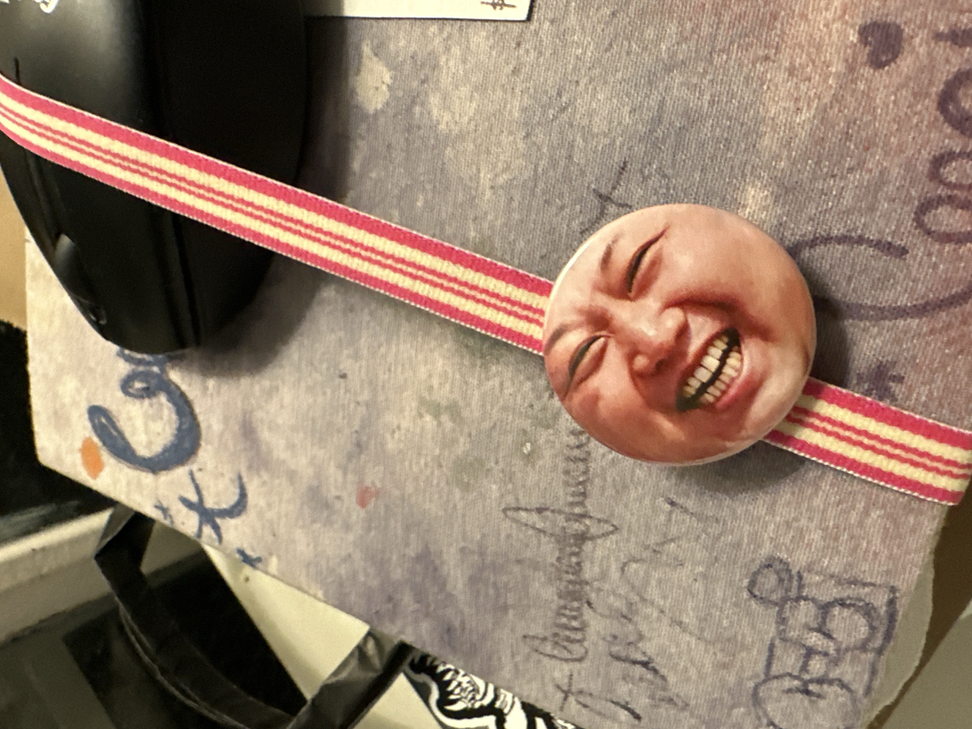 The image shows a close-up view of a book or journal with a textured, greyish-purple cover. The cover has various scribbled markings in blue and dark ink, including what appear to be stylized letters and some abstract shapes. Prominently featured is a pink and cream striped ribbon that runs diagonally across the image. Attached to the ribbon, near the bottom, is a circular pin or button with a close-up image of a person's face. The face is light-skinned with rosy cheeks, eyes closed, and a wide, open-mouthed smile revealing teeth. The background shows a dark object, possibly a computer mouse, and a portion of what looks like a black bag or backpack. The lighting suggests an indoor setting. No specific location in Buenos Aires, Argentina, is identifiable from the image.