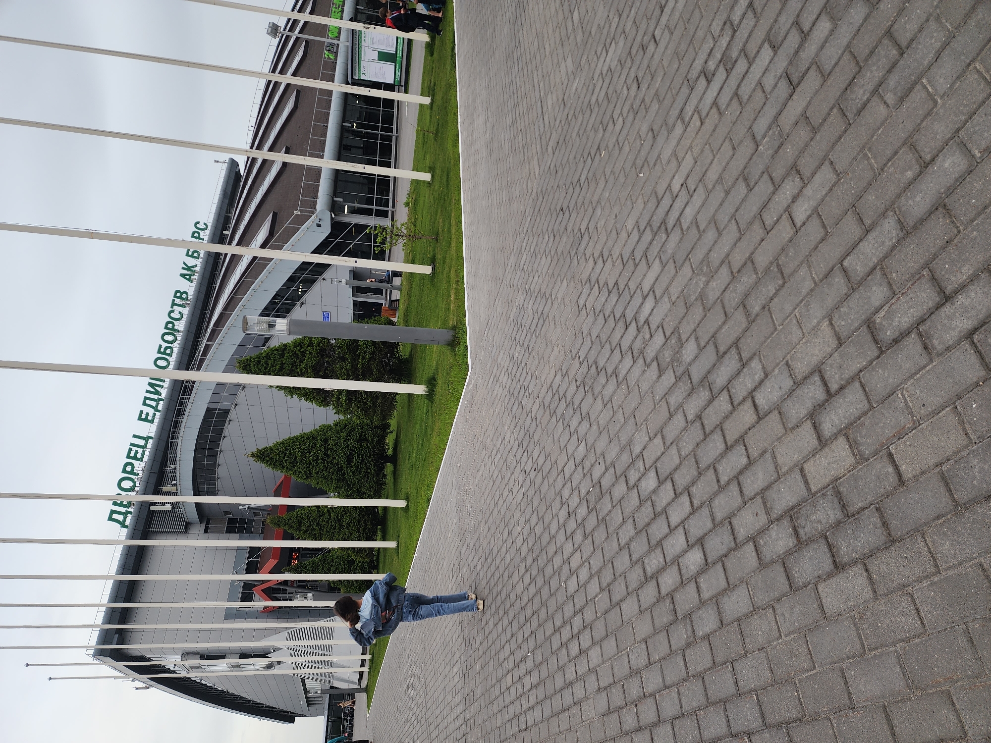 The image shows a young person in jeans and a denim jacket standing on a gray brick pavement in front of a modern building in Kazan, Russia. The building has green lettering that reads "ДВОРЕЦ ЕДИНОБОРСТВ АК БАРС" ("Ak Bars Wrestling Palace"). Tall, white flagpoles are lined up in front of the building. Behind the pavement is a strip of green lawn with small, conical trees. In the background, part of another building with a dark roof can be seen. Some individuals are visible near the second building, likely pedestrians. The sky is overcast. The image has been rotated.