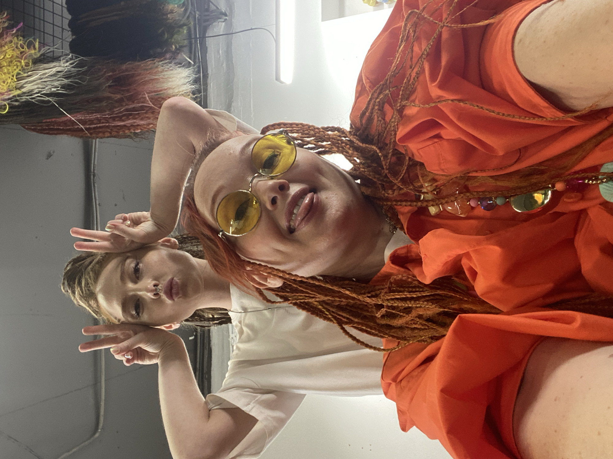 The original image is oriented vertically and rotated 90 degrees clockwise. When viewed upright, the photograph features two women posing playfully in an indoor setting, likely a creative studio or hair salon, in Sochi, Russia.
In the foreground, a woman with prominent reddish-brown dreadlocks and bright yellow circular sunglasses smiles widely, playfully sticking out her tongue. She is wearing a vibrant orange garment, possibly a loose-fitting dress or top, and a long, colorful beaded necklace with various charms around her neck. She appears to be holding the camera for a selfie, looking up at the lens from a low angle.
Behind her and slightly to the left, a second woman with lighter, possibly blonde or light brown, dreadlocks poses with both hands raised, making "peace" signs. She has a nose piercing (septum ring) and a slight pout on her lips, looking directly at the camera. She is wearing a plain, light-colored t-shirt.
The background indicates an indoor location with artificial lighting, including a long white light fixture visible on the ceiling. On the wall behind the women, textured, colorful materials, possibly hair extensions, wigs, or decorative fiber art, are displayed on a black grid structure. The overall mood of the image is casual, friendly, and cheerful. There is no visible text in the image.