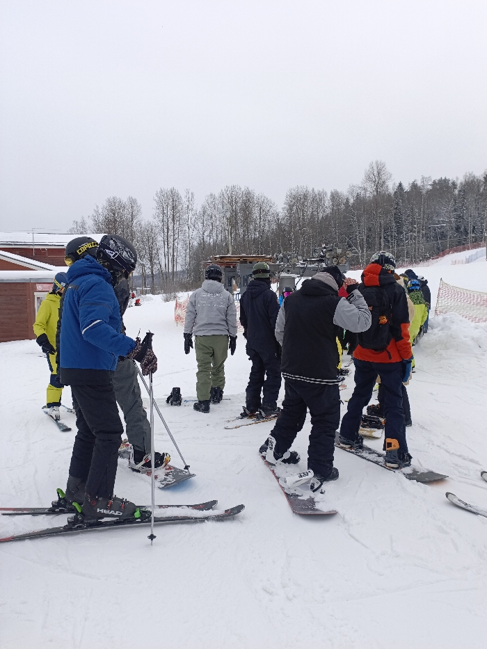 The image depicts a winter sports scene at a ski resort in Svetloe, Russia, under an overcast sky. The entire visible ground is covered in snow. Approximately nine individuals, dressed in winter sports gear, are present, primarily viewed from their backs or sides.
In the foreground left, a person wearing a blue jacket, black pants, a black helmet, and ski goggles stands on skis, holding ski poles. The skis display "HEAD" branding. Adjacent to this individual, another person, largely obscured, is visible in grey pants, equipped with a snowboard. Behind them and slightly to the left, a person in a bright yellow jacket and black pants is also on skis.
The majority of the individuals are positioned in the mid-ground, forming what appears to be a queue. They are equipped with either skis or snowboards. Visible equipment includes various colored jackets (grey, black, red, yellow), helmets, snow pants, ski boots, snowboard boots, and bindings. One individual in a grey hooded sweatshirt and olive-green pants stands on a snowboard. Another, in a black and red jacket with an orange backpack, is also on a snowboard.
In the background, a ski lift mechanism is partially visible. A red-painted building with a white roof is situated on the far left. The horizon features a dense line of bare deciduous trees and evergreen conifers, indicating a forest. Orange safety netting delineates a boundary along the right edge of a snow-covered slope. The scene portrays activity at a winter recreational facility.