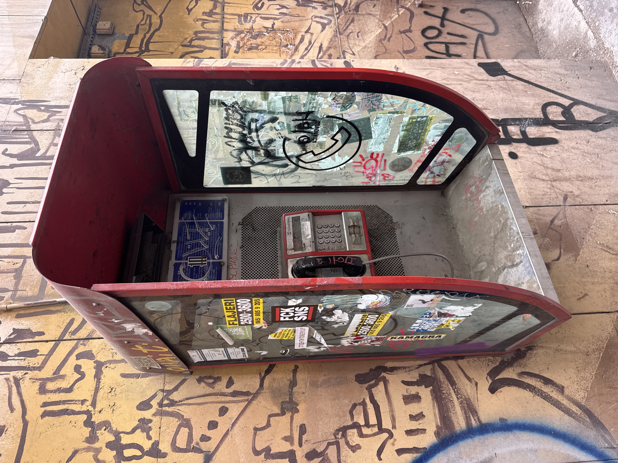 The image shows a red, enclosed, coin-operated public telephone booth attached to a graffiti-covered wall, presumably in Belgrade, Serbia, as indicated by the local telephone numbers and place names on the stickers inside the phone booth. The phone booth itself is weathered and covered in stickers and graffiti on the glass panels. Inside, a red push-button telephone is visible, with a black handset, and blue instruction cards on the left wall. A metal grid platform supports the telephone. The surrounding wall is covered in extensive graffiti, including simple line drawings and tags.