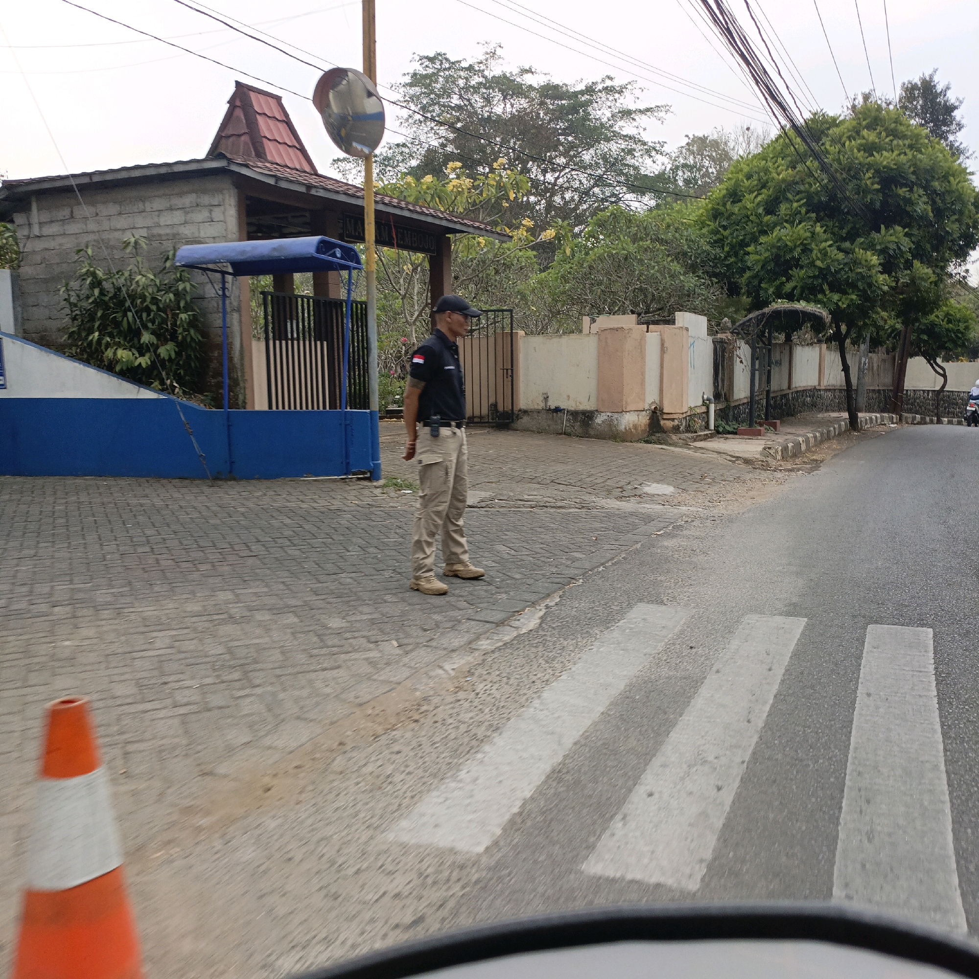 The image depicts a street scene in Salatiga, Indonesia, near the entrance of what appears to be a local government building or educational institution. A man in a dark polo shirt with a white and red armband, khaki pants, and a black baseball cap stands on the pedestrian crossing. He has a radio clipped to his belt and is looking to his left. The building behind him has a distinctive tiered red roof and a gray brick facade, with a blue awning extending from its entrance. A convex mirror is mounted on a pole near the building. The street features a clear pedestrian crossing with white stripes. In the foreground, a blurred orange traffic cone with a white reflective band is visible. The surrounding environment includes trees, a stone wall, and a road with a motorcycle visible in the distance. The sky is overcast.