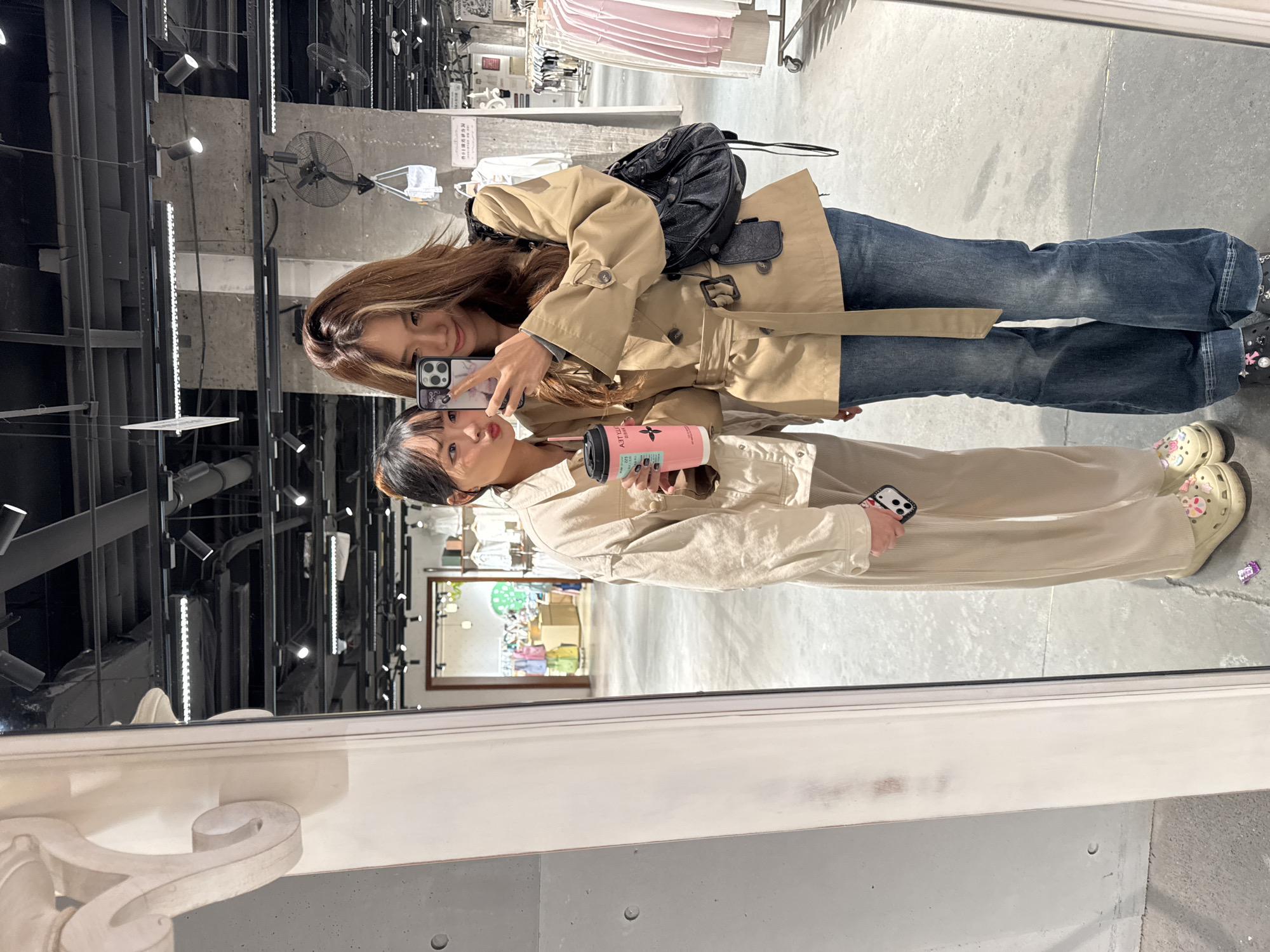 Two women are reflected in a mirror. One woman is wearing a beige trench coat, jeans, and a black backpack, and holding a smartphone. The other woman is wearing beige pants and a beige jacket, holding a pink cup with a straw, and also holding a smartphone. The reflection shows a clothing store interior with clothes racks and lighting fixtures. The mirror is framed by a white, decorative trim. The location is the Jianlan Road District in China, indicated by the Chinese writing on a nearby object.