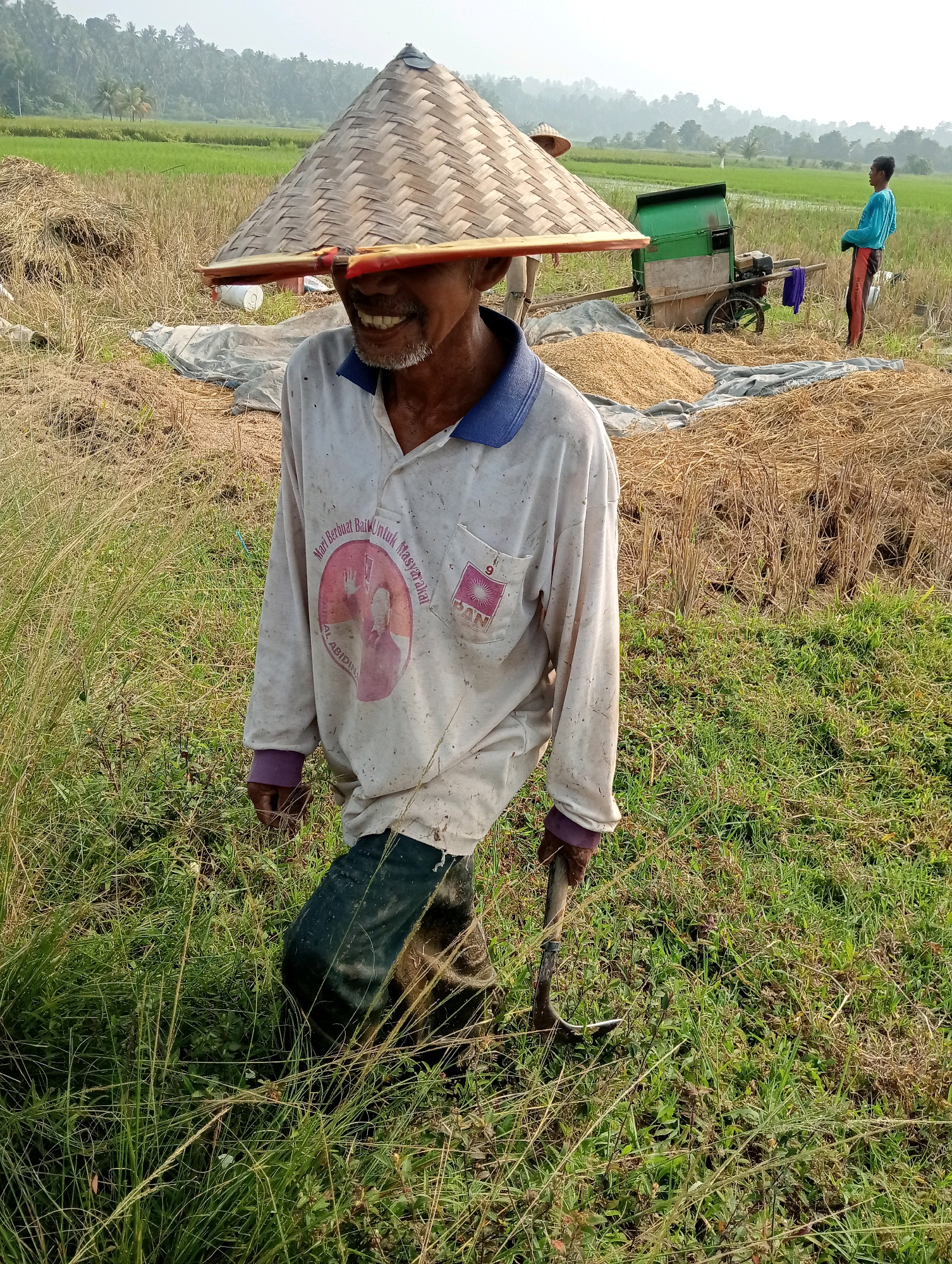 In the city of Campago, Indonesia, a farmer, identifiable by his conical bamboo hat and a light-colored long-sleeved shirt with a faded graphic, is depicted walking through a field. He is holding a shovel in his right hand, its metal blade partially buried in the grass. The man's facial features are visible, including a visible smile and a light beard. The field is a mix of green grass and dried straw, indicative of a recent harvest. In the background, another farmer in a blue shirt stands near a green thresher machine, which is positioned on a tarp spread over straw and rice grains. A pile of harvested rice is visible next to the thresher. Beyond the field, lush green trees form a dense backdrop against a hazy sky. The scene captures a moment of agricultural labor in a rural Indonesian setting.