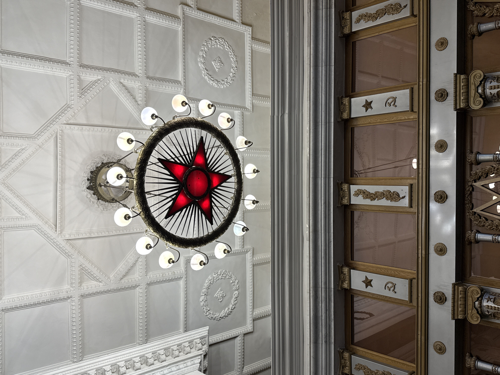 The image shows a view looking up at the ceiling and a portion of the wall of a grand interior space in Moscow, Russia, likely a metro station due to the style of the chandelier and the Soviet-era symbolism.
The ceiling is ornate, featuring a coffered design with intricate decorative moldings and recessed panels. One large central panel is decorated with a circular laurel wreath motif, and other panels have geometric patterns.
A prominent object in the scene is a large, circular chandelier with a red, five-pointed star at its center. The star is surrounded by spokes that extend to the outer rim of the chandelier, from which multiple light bulbs are suspended on curved arms.
Below the ceiling, a section of the wall is visible. It is decorated with a horizontal band of molding, below which are framed panels, likely windows or decorative inserts. Each frame is adorned with columns or pilasters featuring golden laurel leaf designs and Soviet symbols. Specifically, on one of these central pilasters, a red five-pointed star is depicted above a hammer and sickle emblem. Other similar emblems are visible on other pilasters. The windows themselves appear to be tinted a reddish-brown and reflect the ceiling above.
The overall aesthetic is one of grandeur, with a focus on neoclassical and Soviet-era design elements, suggesting a public building such as a Moscow Metro station, possibly like Mayakovskaya or Arbatskaya, known for their elaborate decorations. There are no people visible in the image.