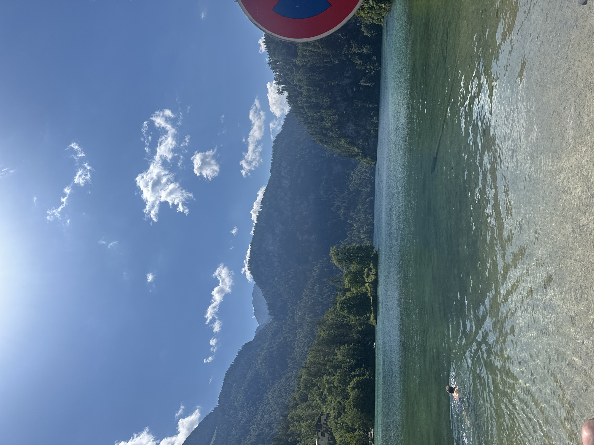 This is an eye-level, medium shot of Königssee lake in Schönau am Königssee, Germany. The sun is shining brightly, casting light across the calm, clear water, which reveals the rocky lakebed in the foreground. A person is swimming in the lake, facing away from the camera. The background features a densely forested mountain range that rises steeply from the lake's edge. A small island with a single tree is visible in the middle distance. On the right side of the frame, a red and blue "no parking" sign is partially visible. The sky is a clear blue with scattered white clouds.