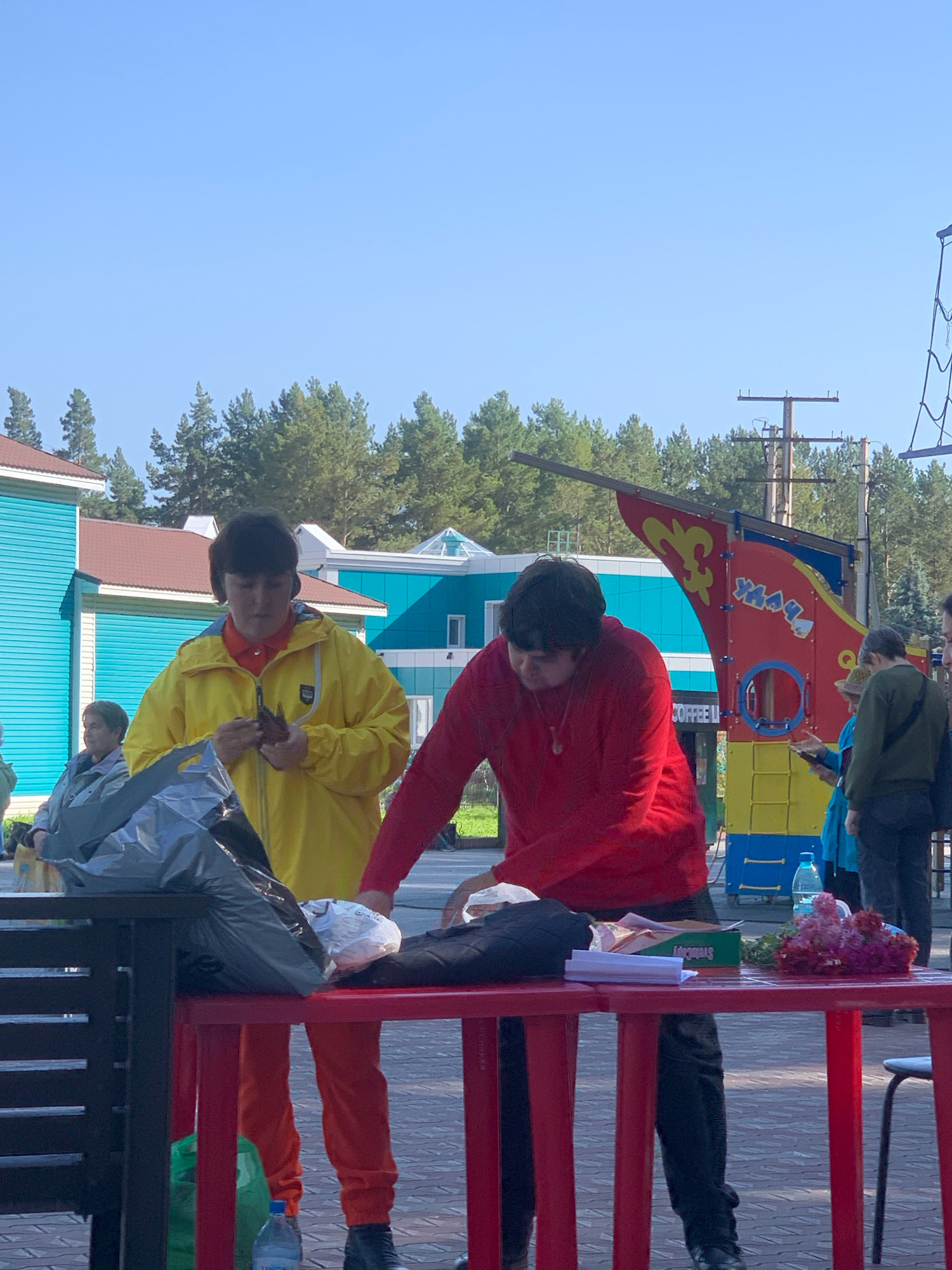This image captures an outdoor scene, likely in Prokopyevsk, Russia, given the context. Two individuals are the primary focus, standing behind a red table. One person, wearing a yellow jacket and orange pants, is examining a small, dark object in their hands. Adjacent to them, another individual in a red long-sleeved shirt is leaning over the table, appearing to handle items.
The table itself holds several objects, including a bouquet of purple and pink flowers, a green box, a stack of white papers, and plastic bags, one of which is dark and crinkled. A partially visible water bottle with a blue label sits on the ground near the table. In the background, a vibrant, red and yellow playground structure resembling a ship's prow is visible, along with buildings featuring turquoise and brown facades. Trees line the horizon, and a clear blue sky dominates the upper portion of the image. Other people are partially visible in the background, suggesting a public gathering or event.