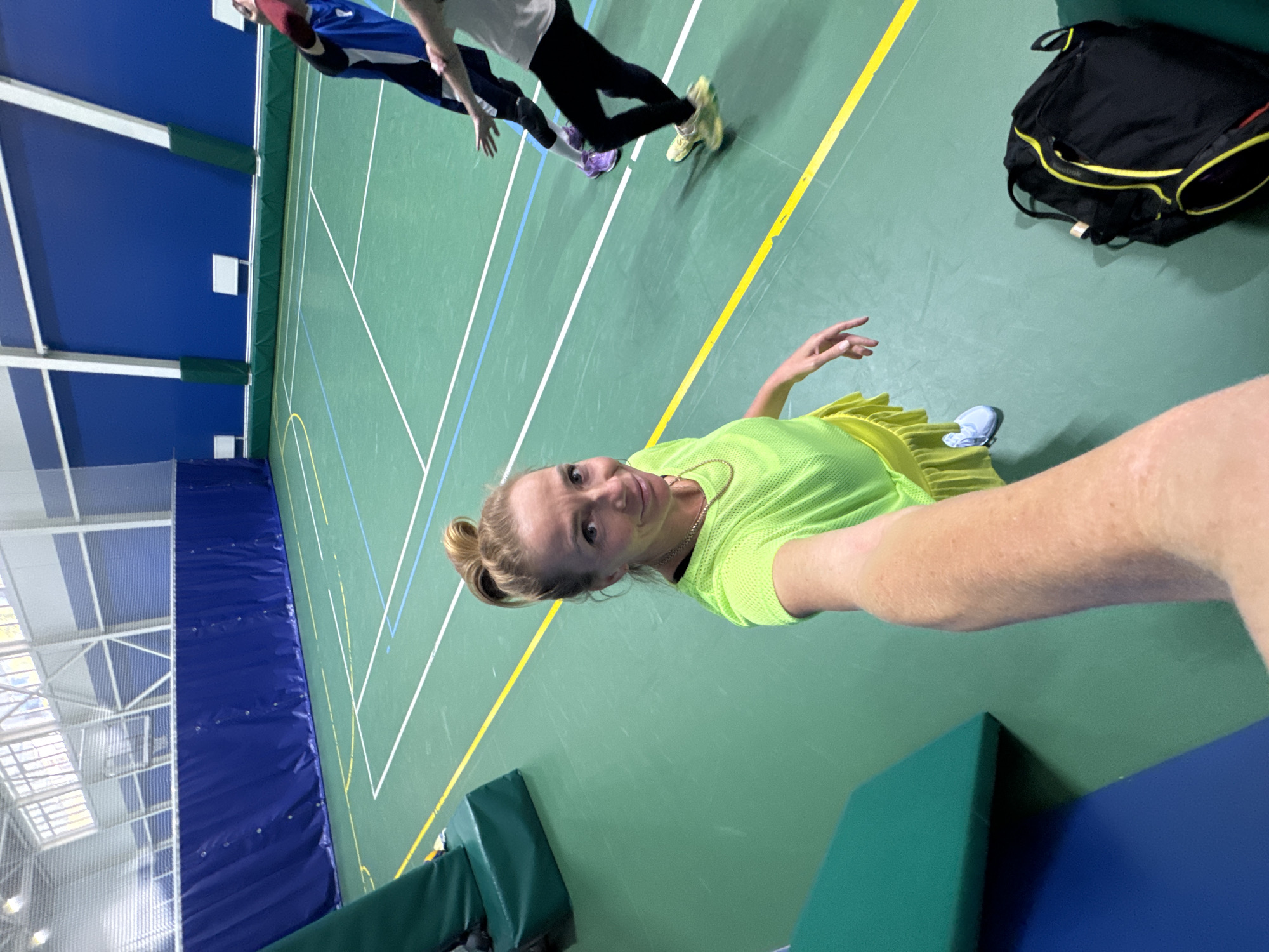 The image shows the interior of a sports hall. A woman is taking a selfie, wearing a bright green sports top. Two other people are visible in the background. One person is wearing a blue and white top, while the other is wearing a grey top with black leggings. Lines are painted on the green floor, indicating sports court markings. A black and yellow sports bag is on the floor. A blue padded wall is on the left. The location is a sports hall, likely in Saint Petersburg, Russia.