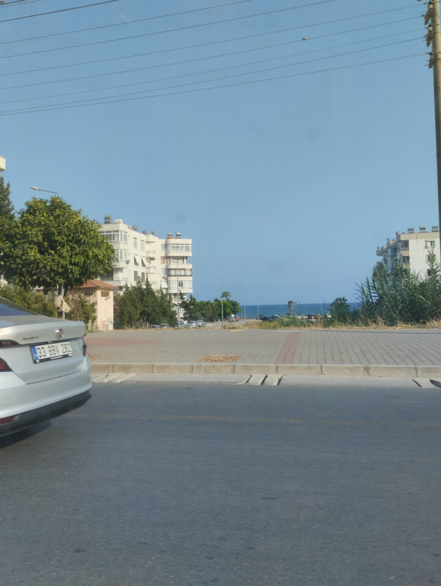 In Mezitli, Turkey, a silver sedan is parked on the left side of the frame, its rear visible, facing away from the viewer. The car's license plate displays the characters "33 BBV 282". Beyond the car, a paved area with interlocking concrete blocks stretches towards a row of buildings. The buildings are modern, multi-story structures with white facades and numerous windows. Some buildings feature solar water heaters on their roofs. To the left, a lush green tree with a thick canopy stands near a small, tan, shed-like structure. The background reveals a glimpse of the Mediterranean Sea under a clear blue sky, with a few distant palm trees dotting the coastline. Power lines crisscross the sky above the scene. The overall environment suggests a sunny day in a coastal urban area.