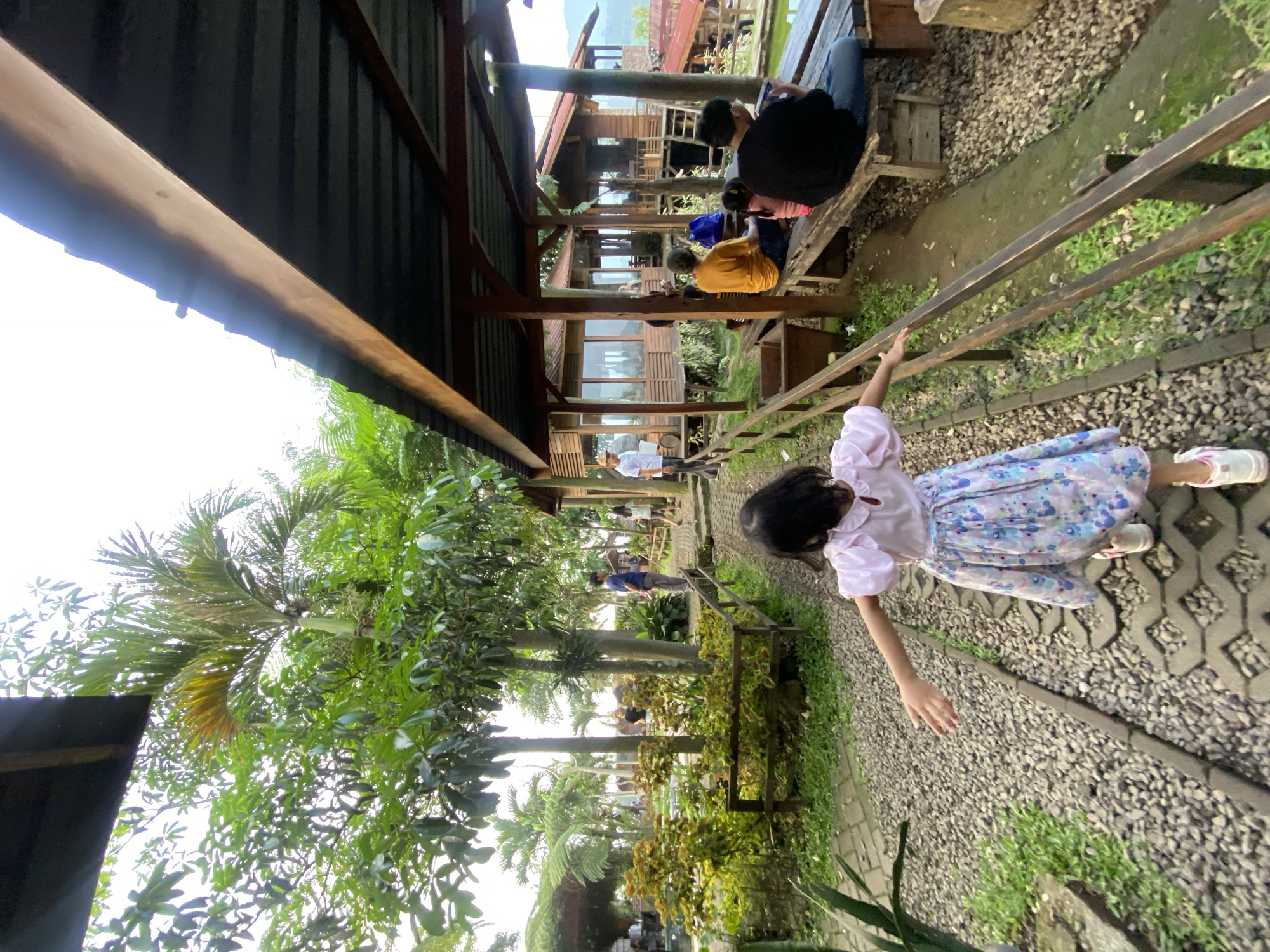 The image, taken in Selotapak, Indonesia, shows a young girl in a pink blouse and floral skirt walking along a path with a wooden railing. The path appears to be made of gravel and pavers. The girl's left hand is extended toward the railing. In the background, people are seated on benches under a covered structure, suggesting an outdoor cafe or rest area. Lush green vegetation, including palm trees, surrounds the area. Buildings can be seen further in the distance, indicating a populated area. The scene suggests a leisurely setting within a semi-urban environment.