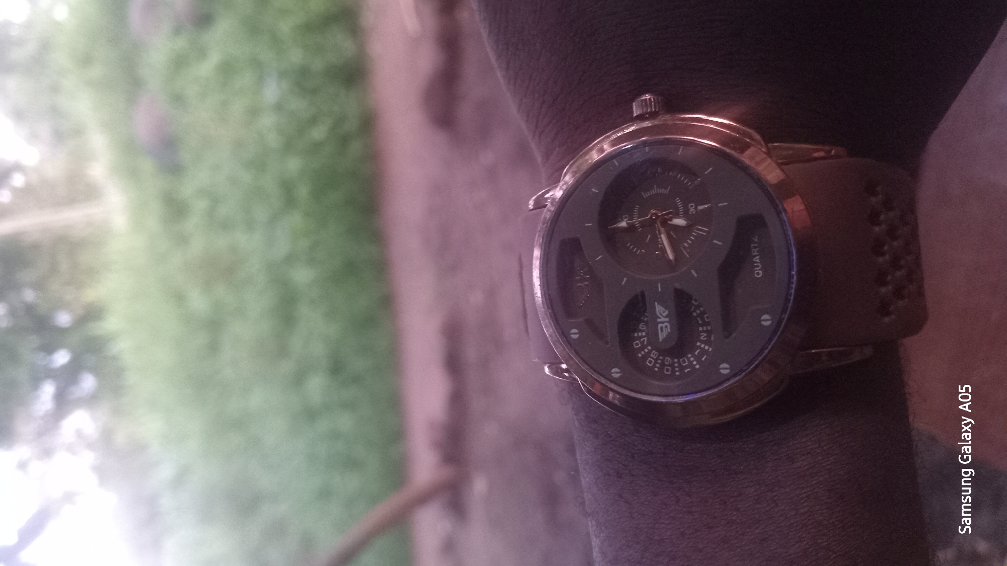 A close-up, eye-level shot focuses on a dark-faced wristwatch with a brown band worn on a person's wrist. The watch face features multiple sub-dials and hands, with the word "QUARTZ" visible. The background is blurred, showing lush green foliage and possibly some trees, suggesting an outdoor setting in Ribina East, Nigeria. The lighting appears somewhat overcast. The bottom left corner of the image displays "Samsung Galaxy A05".