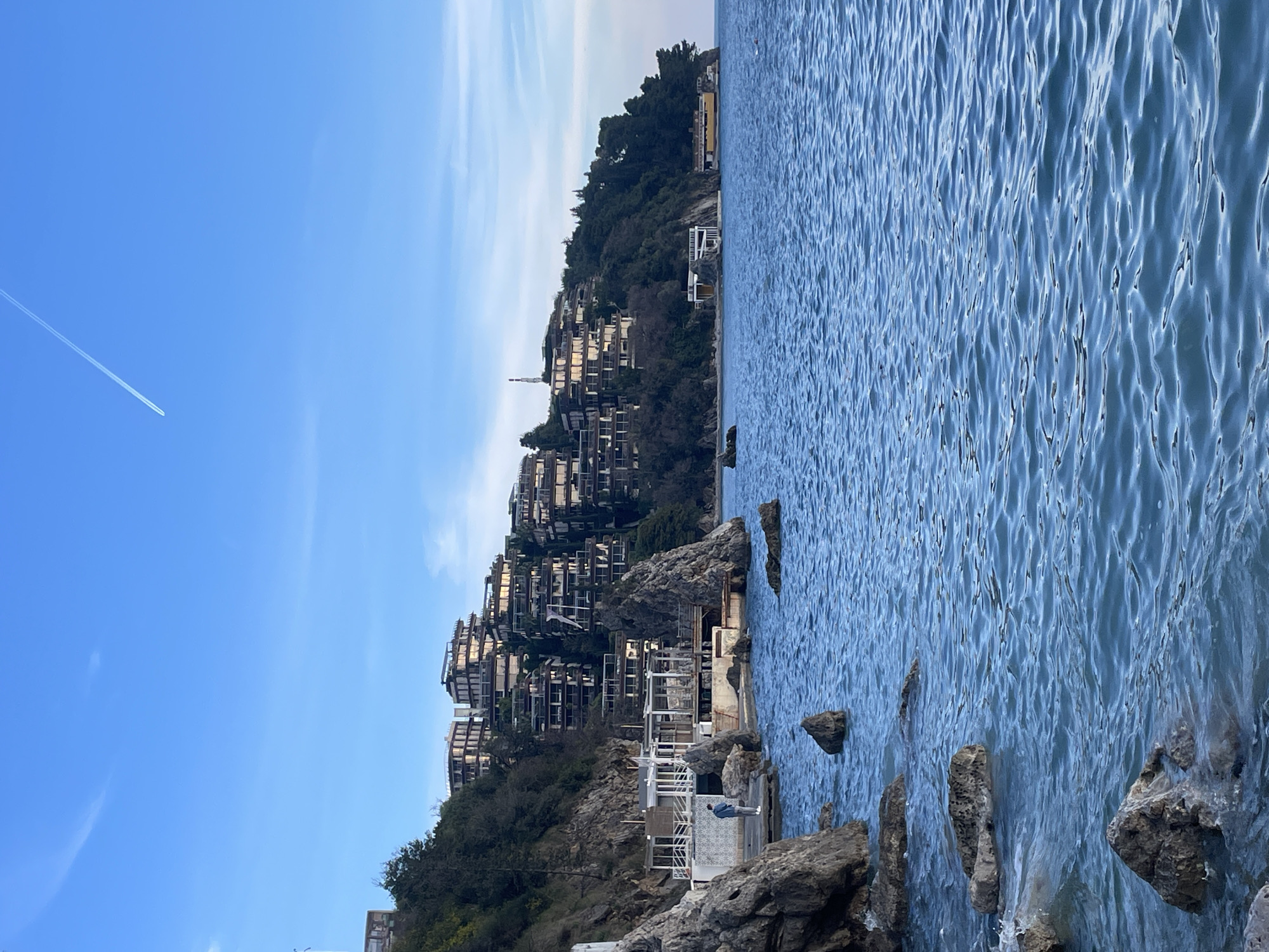 This image depicts a coastal scene in Budva, Montenegro, specifically overlooking the Zavala Peninsula, featuring the Dukley Gardens residential complex.
The environment is a clear, daylight outdoor setting. The sky is light blue with a single, linear white contrail from an aircraft extending diagonally across the upper left quadrant. The sea, occupying the lower half of the frame, is a calm, blue-green body of water with gentle ripples across its surface.
In the foreground and left midground, a rugged shoreline is present, characterized by numerous dark gray boulders, some partially submerged by the water where small waves are breaking. Directly on the coastline, a multi-level white building complex, likely a beach club or restaurant, is constructed on the rocks. This complex features large windows and white railings.
One person is visible on a concrete platform adjacent to the white building complex, facing the water. This individual is dressed in a light blue jacket, light-colored pants or jeans, and white shoes.
The background is dominated by a steep, tree-covered hillside rising from the coast. Perched on this hillside are numerous multi-story, terraced residential buildings belonging to the Dukley Gardens complex. These structures display modern architecture with large balconies and reflective glass facades. A slender white communication mast or tower is visible on the hill's summit, situated slightly to the right of the main cluster of buildings. To the far right, the tree-covered hillside continues, and another smaller white structure with a yellow section is visible at the waterline.