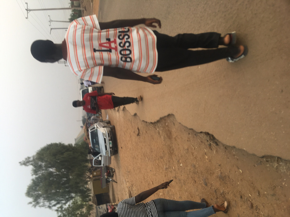 This image captures a street scene in Pindiga, Nigeria, featuring several individuals on or beside a road. The perspective is from slightly behind and to the right of the primary subject, looking down the road.

In the immediate foreground, a person is partially visible on the right, seen from the back, wearing a striped top and blue jeans, holding a mobile phone in their right hand.

The central figure is a young man, also seen from the back, walking away from the camera on the road. He wears a white short-sleeved t-shirt with thin red horizontal stripes and black pants. On the back of his shirt, prominent red and black lettering reads "1A BOSSE". He is wearing open-toed sandals and appears to be holding a small dark object, possibly a phone, in his left hand.

Further down the road, another young man, also viewed from the back, is walking. He wears a solid red t-shirt and dark pants, also in sandals.

The road itself appears to be partially paved, with significant dirt and erosion along its edges, indicating a potentially uneven or rural pathway. A white pickup truck is parked on the left side of the road, with its driver's side door open. A motorcycle is parked near the truck. In the background, a large tree with green foliage is visible on the left, and utility poles with power lines stretch across the sky. The sky is uniformly light, suggesting either an overcast day or hazy conditions.