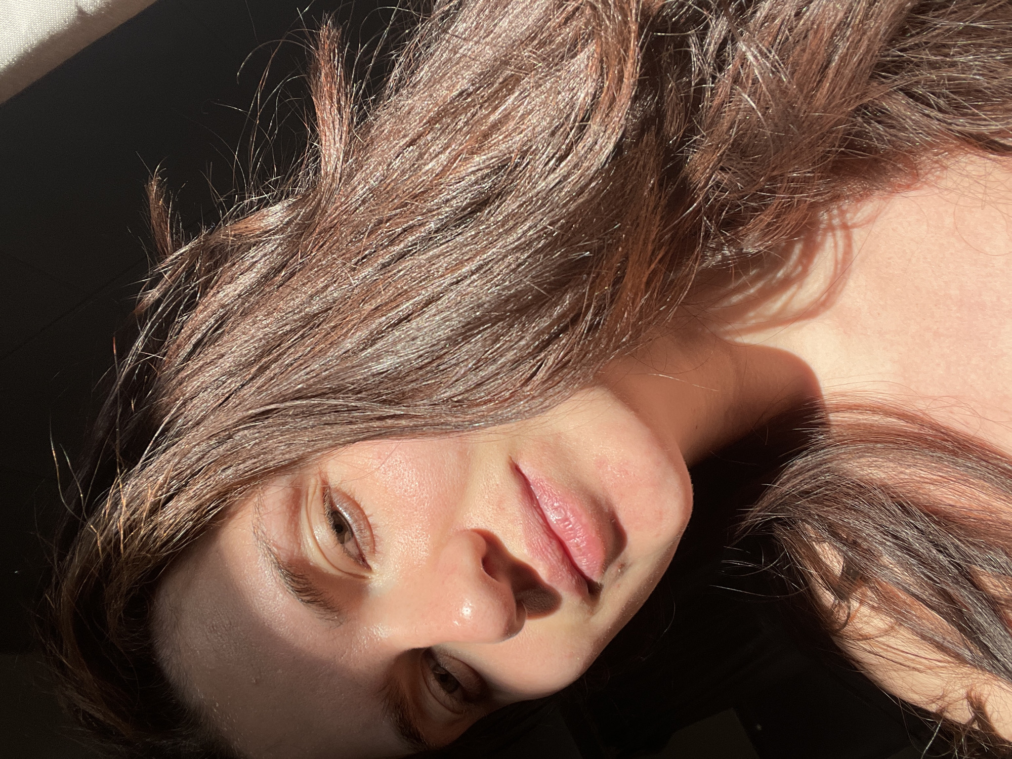 A close-up, angled portrait depicts a woman with long, light brown hair. Her face is partially illuminated by sunlight, highlighting her skin texture. Her eyes are visible, and she has a fair complexion. The background appears dark and indistinct. There is no indication of location in Мосметаллоконструкция Онт, Russia.