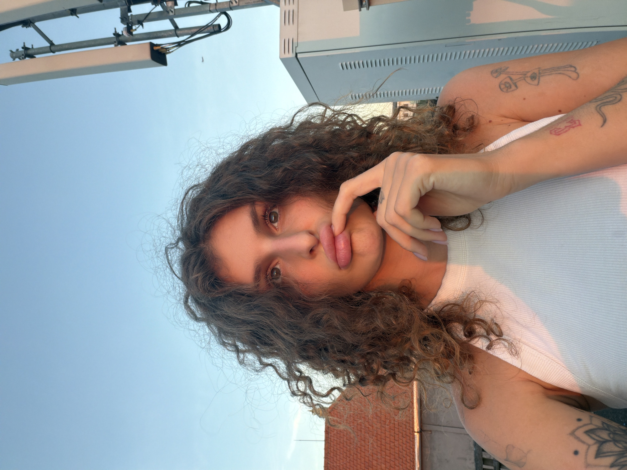 A woman with curly brown hair and light skin is positioned in front of an urban backdrop. She is wearing a white tank top and has tattoos on her arms. One hand is near her face, touching her lips. Above and behind her are metallic structures and a light-colored building. The background includes a blue sky and glimpses of rooftops with terracotta tiles. Based on the available information, the exact location in North Kuta, Indonesia cannot be determined definitively.