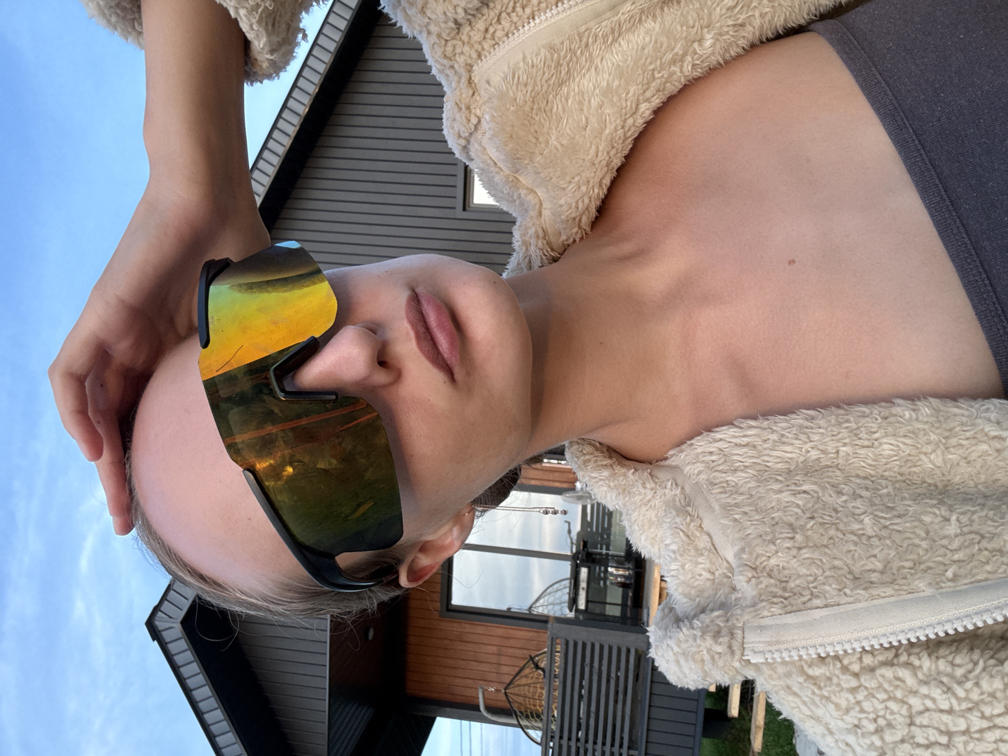 The image shows a young woman taking a selfie outdoors. She is wearing large, reflective sunglasses and a fluffy cream-colored jacket over a dark grey top. Her right hand is raised to her forehead, and she is looking towards the camera. In the background, there is a modern, wooden house with a dark grey sloped roof, and some outdoor furniture, including a hanging chair. The sky is a pale blue with hints of clouds. The image appears to have been taken during daylight hours. The location is not specified as Podshivalovo, Russia.