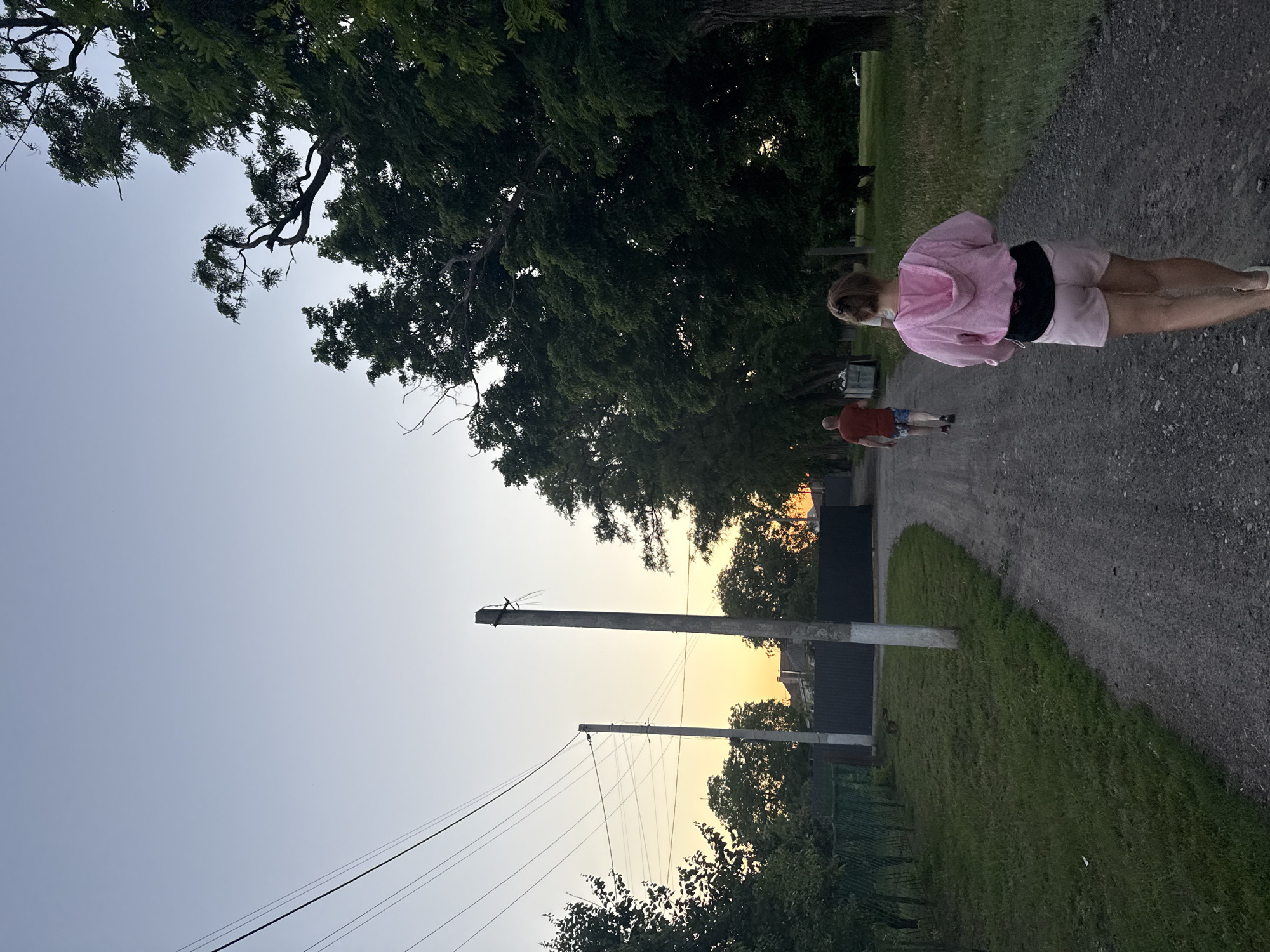 This image depicts a rural scene in Obukhivka, Ukraine, during twilight. Two individuals are walking away from the viewer on a gravel road. The person in the foreground is wearing a pink hooded jacket and light pink shorts, with their back to the camera. Further down the road, a person in a red t-shirt and patterned shorts is walking ahead. The road is bordered by a grassy area on the left and a line of tall trees on the right. Utility poles with power lines are visible in the background, along with some residential structures and a dark fence. The sky is a pale gradient from light blue at the top to a soft orange hue near the horizon, indicating sunset or sunrise. The overall atmosphere is peaceful and suggests a casual outdoor activity.
