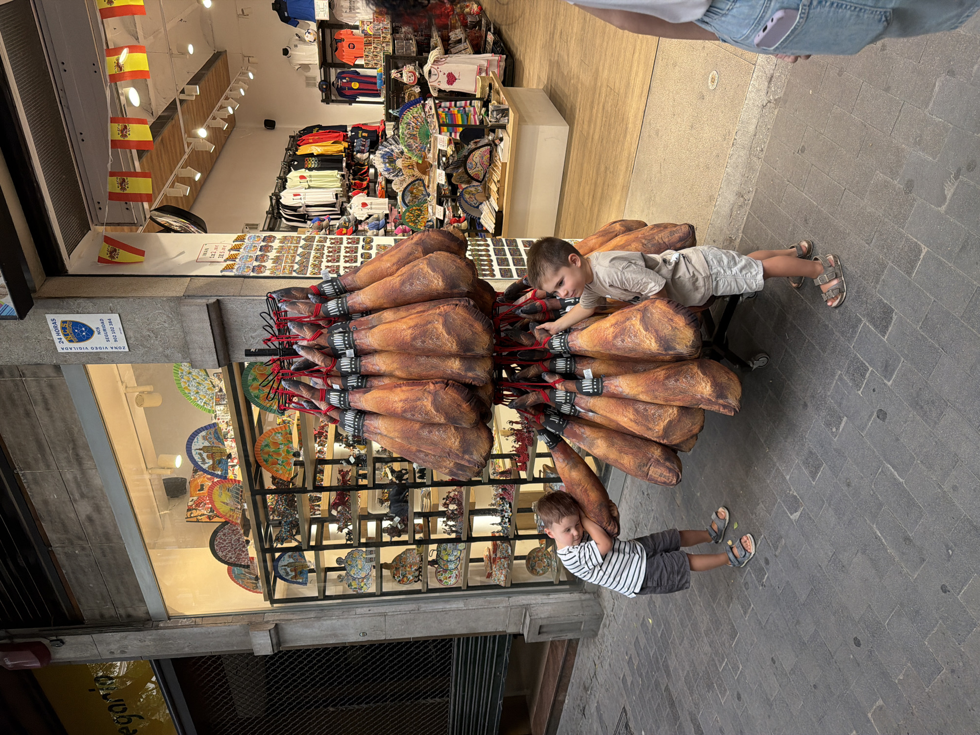 Two young boys are in front of a shop that sells Spanish hams and souvenirs. The boy on the left, wearing a striped t-shirt and shorts, is leaning on a large cured ham, with his eyes closed. The boy on the right, wearing a light-colored t-shirt and shorts, is reaching out to touch one of the hams. The shop window displays numerous colorful fans and ceramic figures, including bulls and flamenco dancers. Flags of Spain are hanging above the shop entrance. The ground is paved with dark stone tiles. The overall scene appears to be a lively street in a tourist area.