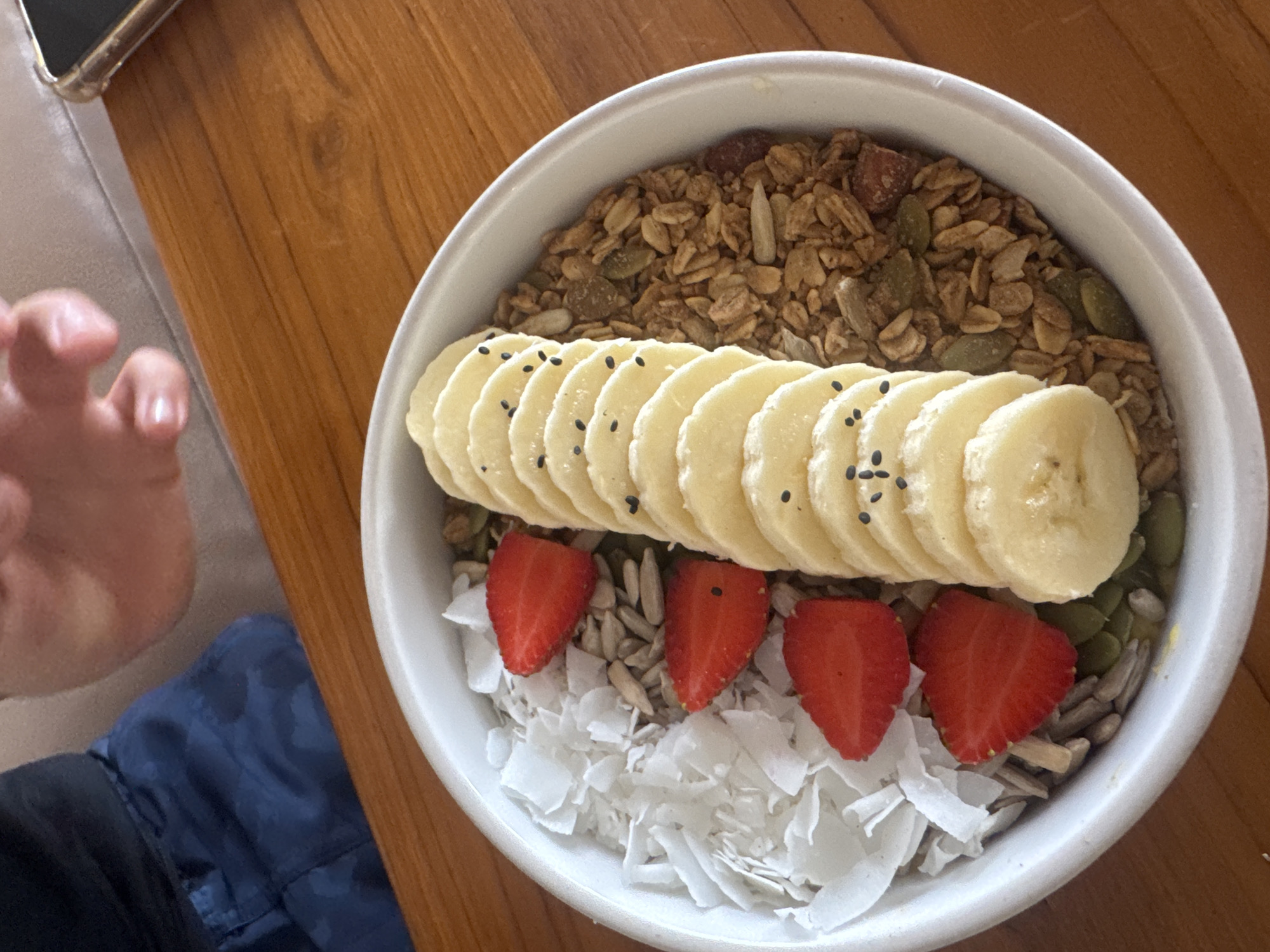 A white bowl of acai or smoothie bowl sits on a wooden table. The bowl is sectioned into three main parts. On the left, it is filled with shredded coconut and sunflower seeds. In the middle, slices of banana are arranged vertically, sprinkled with black sesame seeds. On the right, the bowl is filled with granola and pumpkin seeds. Three sliced strawberries are placed on top of the shredded coconut. A person's hand with a black shirt and blue camouflage shorts is visible in the upper left of the frame, and a portion of a phone case is visible in the upper right. The scene is in Kuta Selatan, Indonesia.