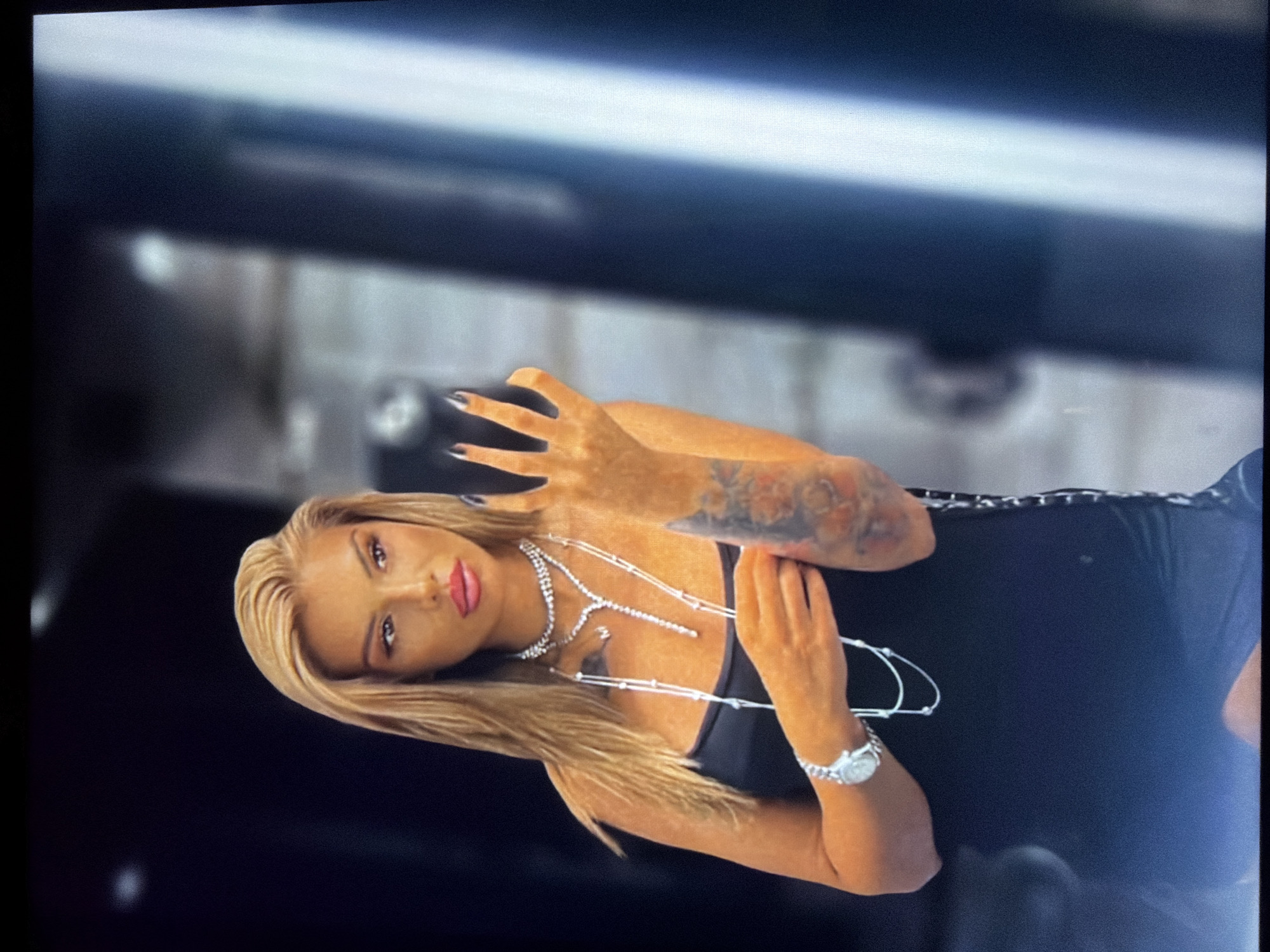 A woman with long, straight blonde hair and tanned skin is depicted in a portrait-style shot, rotated 90 degrees clockwise. She is wearing a black top or dress with a lace-up detail visible on her right side. Her arms are adorned with tattoos, and she has long, dark-colored nails. She wears several pieces of jewelry, including multiple sparkling necklaces, one of which is a choker, and a prominent silver-toned watch on her left wrist.
Her facial features include full, dark pink lips and defined eyebrows, with her gaze directed towards the viewer. She is holding a dark-colored smartphone in her right hand, positioned near her face. Her left hand is resting near her chest, gently touching the necklaces.
The background is dark and heavily blurred, suggesting an indoor setting with low light or a studio environment designed to keep the focus on the subject. There are subtle hints of lighter, possibly reflective, surfaces in the background, but no clear identifying features. The overall image appears to be a capture from a screen, exhibiting some pixelation and reflections, particularly noticeable in the upper left corner. No text is visible in the image.