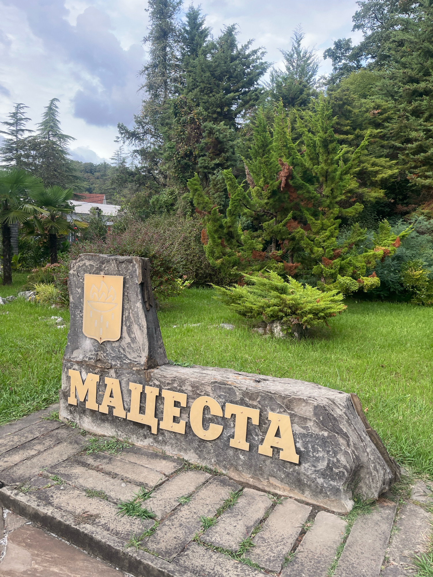 The image depicts a sign made of a large, rough stone with the word "МАЦЕСТА" (Matsesta) inscribed in gold Cyrillic letters. Above the inscription, a gold shield with a stylized emblem is affixed to the stone. The sign is set on a stone pathway, surrounded by a well-maintained lawn. In the background, lush green trees and foliage create a natural setting. The sky is overcast with clouds. The location is Matsesta, a district in the city of Sochi, Russia, known for its mineral springs and health resorts. There are no people visible in the image.