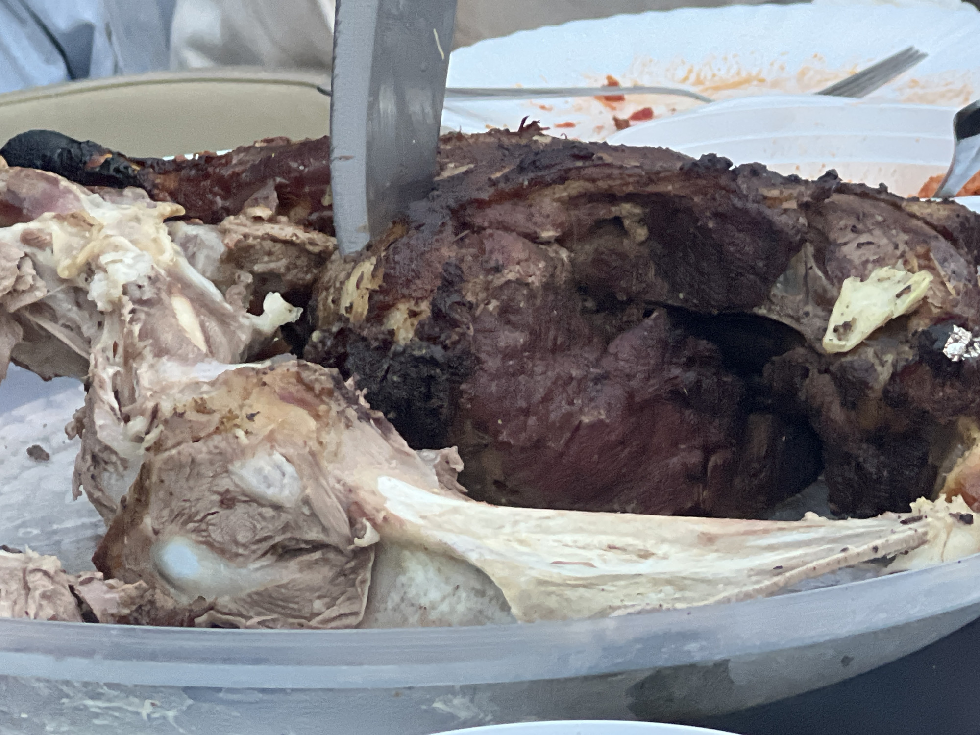 A close-up shot captures a whole roasted animal carcass, likely a lamb or goat, in a clear plastic container. A metal knife is inserted into the roasted meat, suggesting it is being carved or served. The meat is dark brown and appears well-cooked, with some visible bones and sinew. The background is blurred, but hints of white tablecloths and plates suggest an outdoor or informal dining setting. No specific location within the city of Bender, Moldova, can be determined from the image.