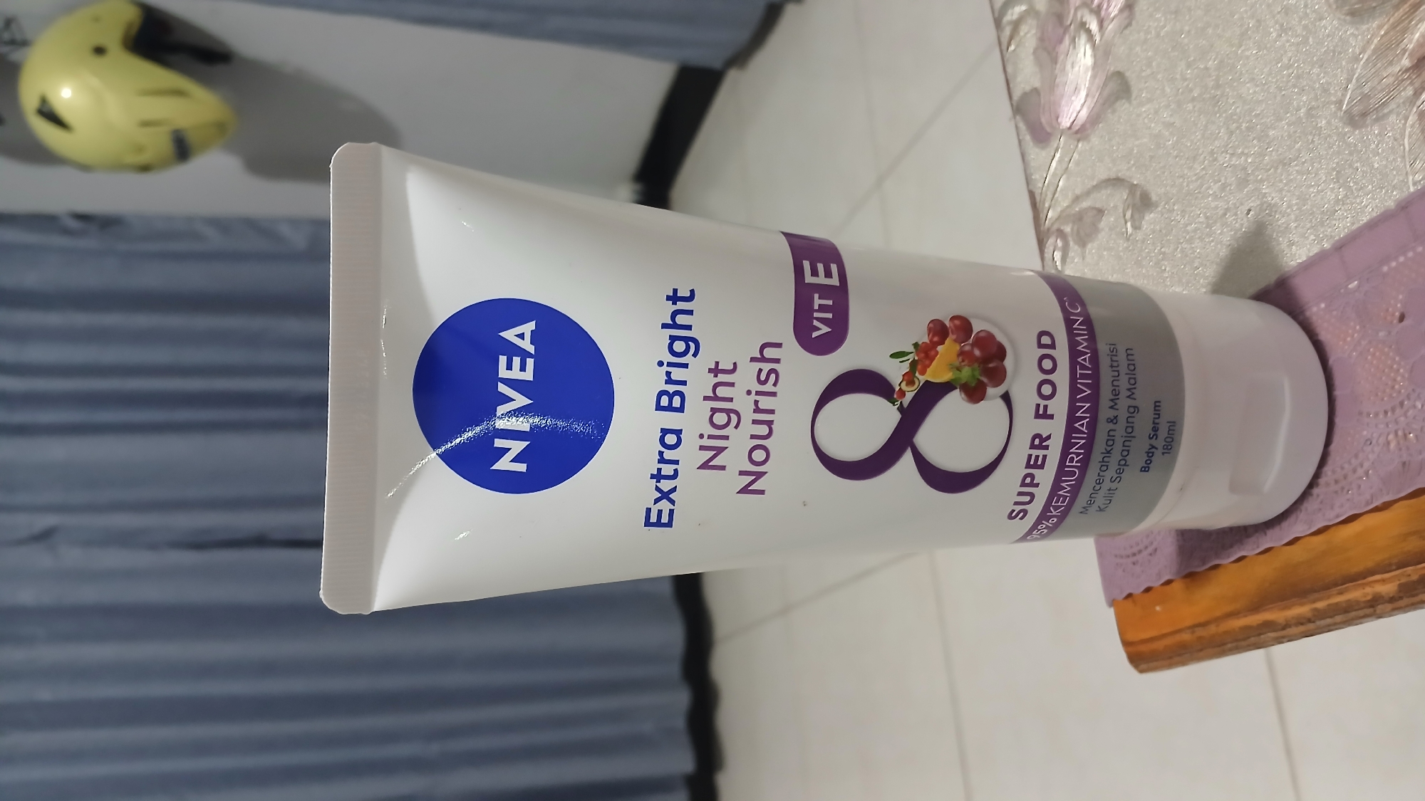 A white Nivea Extra Bright Night Nourish body serum tube is positioned upright on a wooden surface with a textured fabric underneath. The tube features the Nivea logo in blue, followed by the product name in purple text. Below this, a large purple number '8' is displayed, accompanied by an illustration of grapes, berries, and what appears to be a slice of orange or mango. Further down, text indicates "SUPER FOOD" and "95% KEMURNIAN VITAMIN C" (95% Vitamin C Purity) in Indonesian. The bottom of the tube reads "Mencerahkan & Menutrisi Kulit Sepanjang Malam" (Brightens & Nourishes Skin All Night Long), "Body Serum", and "180ml". In the background, a gray curtain hangs behind the product, and to the right, a yellow helmet is mounted on a white wall. The floor beneath the wooden surface appears to be tiled. This scene is consistent with a product display or personal use setting within a home or store in Sampang, Indonesia, given the Indonesian language on the product packaging.