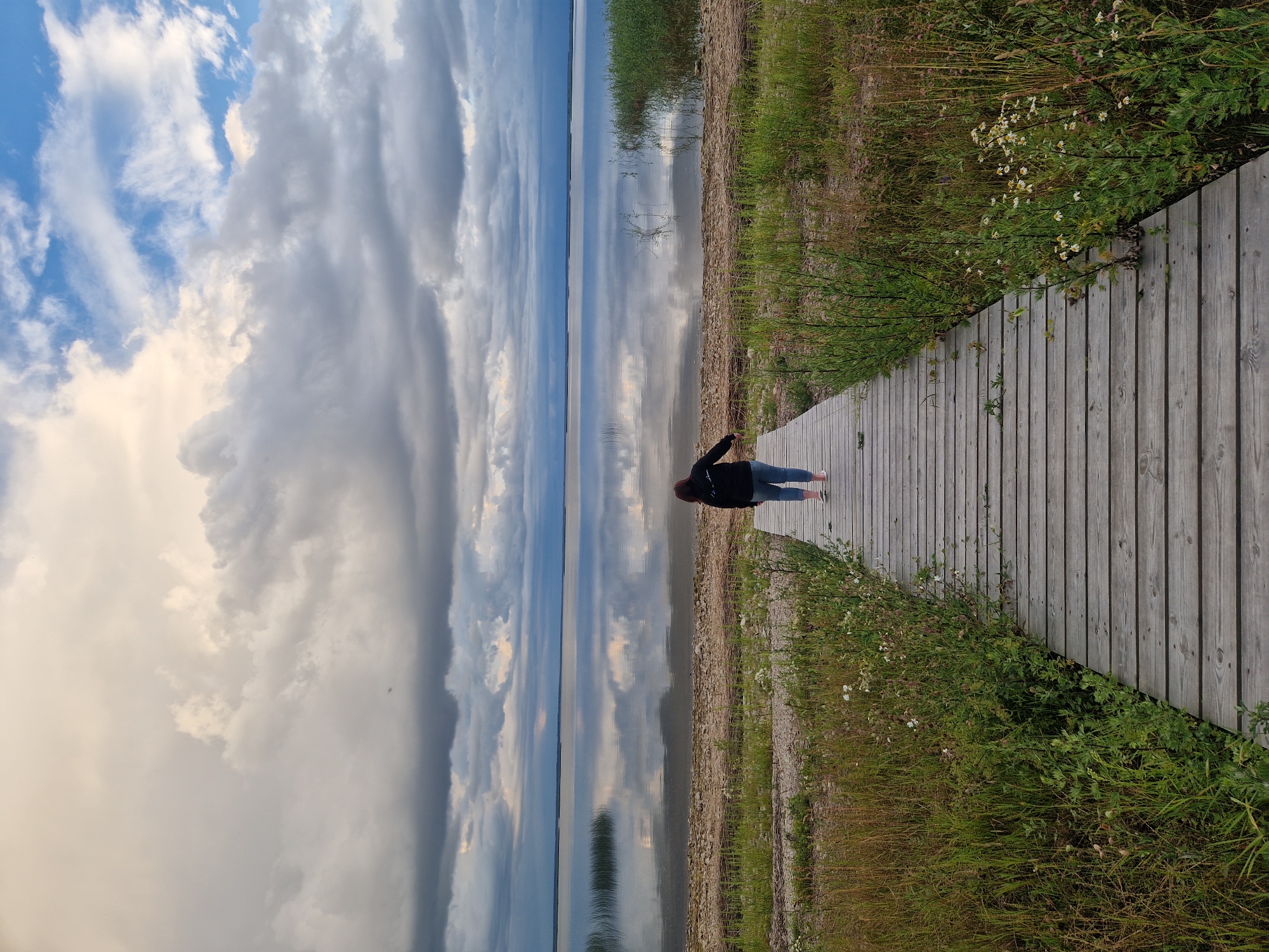 A person stands on a wooden boardwalk, facing a calm body of water. The boardwalk is made of weathered gray planks and runs diagonally across the right side of the frame, bordered by tall green grass and some wildflowers. The water reflects the dramatic sky, which is filled with a mix of bright white and dark gray clouds, suggesting an overcast or rapidly changing weather day. The horizon line is clearly visible where the water meets the sky. The person is wearing a dark jacket and light-colored pants. Their pose, with one arm slightly raised, suggests contemplation or enjoyment of the scenery. The overall impression is one of peace and connection with nature. The time of day appears to be late afternoon or early evening, given the quality of light. The location is identified as Īdeņa, Latvia. There is no visible text in the image.