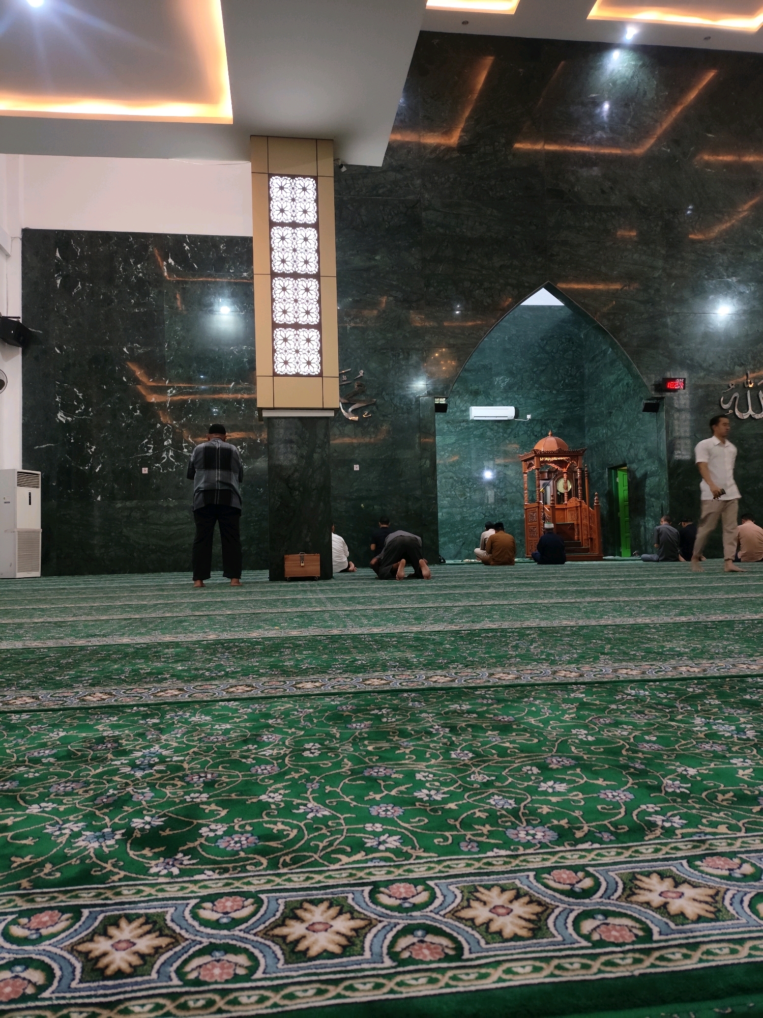 This media file captures an interior view of a mosque in Palembang, Indonesia. The scene depicts several individuals engaged in prayer, with some in standing positions and others in a kneeling posture on a richly patterned green carpet. The mosque's architecture features walls clad in dark green marble, adorned with embedded linear lighting that creates a dramatic effect.
In the foreground, the intricate floral design of the mosque's carpet is prominently displayed. Further back, a man stands facing away from the camera, dressed in a checkered shirt and dark pants, appearing to be in prayer. To his left, an air conditioning unit is visible. Towards the center of the image, a wooden minbar, a pulpit used for sermons, stands within an arched alcove. Several other individuals are seated or kneeling in prayer on the carpet, facing the minbar and the qibla wall.
Above the minbar, a digital clock displays the time as 18:29, suggesting it is late afternoon or early evening. Arabic calligraphy, likely the word "Allah," is visible on the wall to the right of the clock. The ceiling is modern, with recessed lighting providing a warm illumination. The overall atmosphere appears serene and devotional, indicative of prayer time within the mosque. The presence of the minbar and the directional lighting on the walls suggests this is a place of worship and reflection.