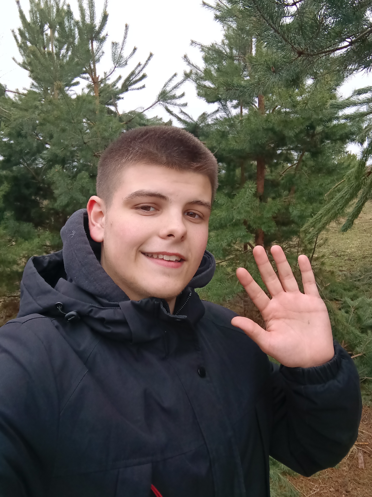 A medium-shot photograph shows a young man posing in an outdoor setting. He is standing among pine trees, which are green and lush. The man is fair-skinned, with short, light-brown hair. He is wearing a black hooded jacket. He is smiling and waving at the camera with his right hand. The background features other trees and foliage, suggesting a wooded or park-like area. The location is the city of Krasnyi Mayak, Russia.