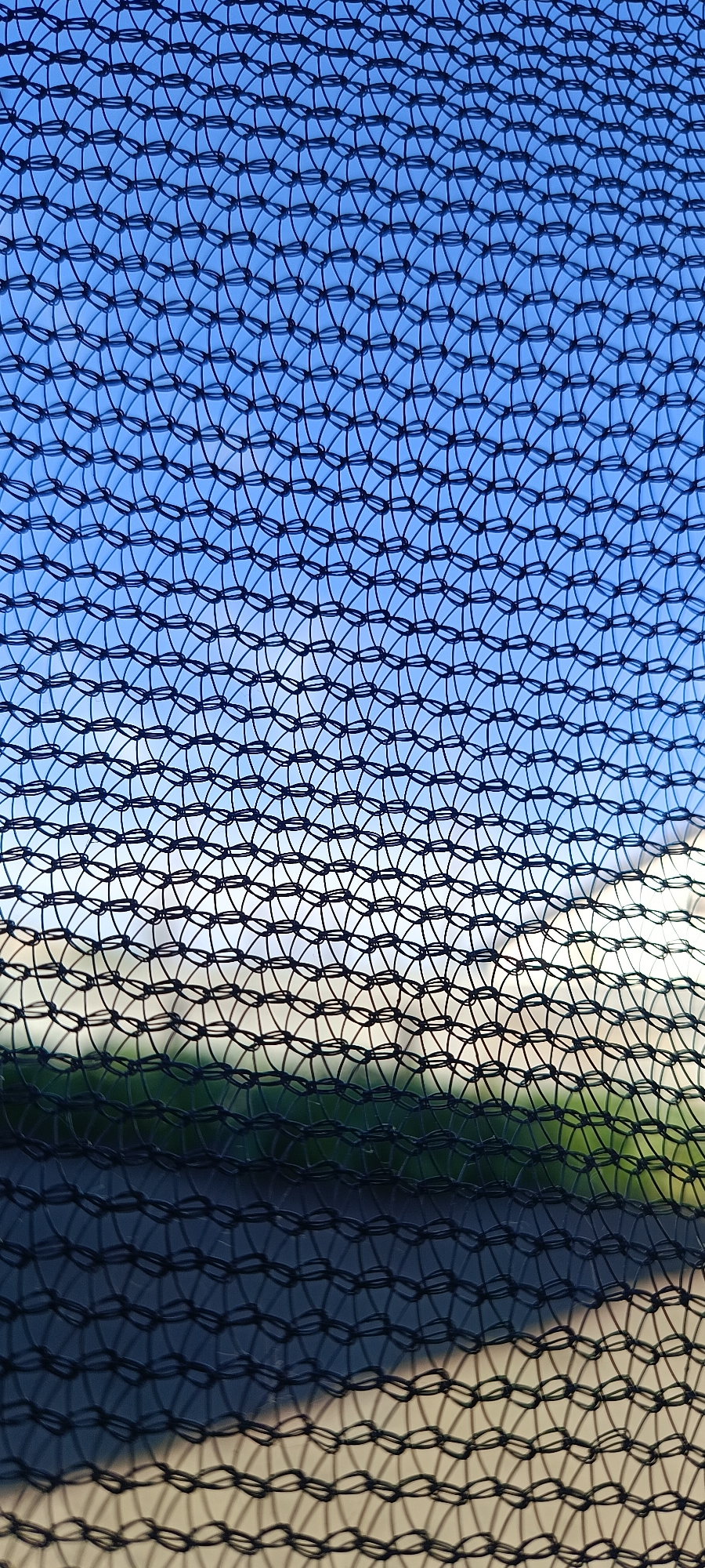 The image is a close-up shot of a black net with a blue sky and a hint of green landscape visible through it. The net creates a repetitive pattern of interconnected loops, obscuring the view of the background. The sky is clear and bright, suggesting it is daytime with good weather. The lower portion of the image shows a blurred area of green, likely grass or trees, and a lighter, possibly sandy or paved, surface. Shadows are cast by the net onto the ground below, adding depth to the scene. There is no discernible text or activity taking place. The overall impression is abstract due to the dominant pattern of the net.