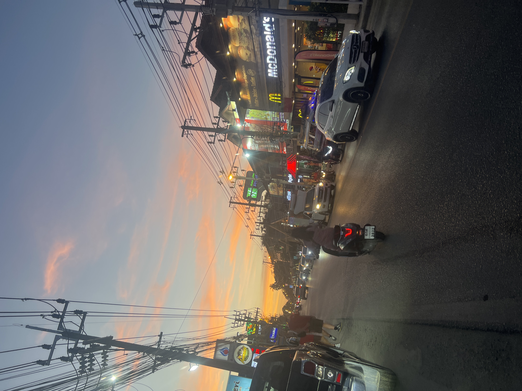 This image depicts a street scene in the city of Ko Samui District, Thailand, during sunset. The sky displays a gradient of warm colors, from soft oranges and pinks near the horizon to a pale blue overhead, with scattered clouds. A complex network of utility poles and overhead wires dominates the left and upper portions of the frame, obscuring much of the sky.
The street is lined with buildings housing various businesses. Prominently visible is a McDonald's restaurant with its signage lit up. Other establishments include what appears to be a "Holiday Inn Resort" with green signage and an establishment with a red awning and a "Pizza Hot" sign. Signs for "Taco Bell" and "Good Times" are also discernible.
Traffic occupies the street, with several vehicles including cars and motorcycles. A white Mercedes-Benz SUV is in the foreground on the right. On the left side of the street, a black pickup truck is parked or moving slowly. A person is riding a dark-colored scooter in the middle of the road, with its taillight illuminated and a license plate visible. Two individuals, one wearing a red shirt and shorts, and another in a red shirt and darker shorts, are walking on the sidewalk on the left side of the street.
The road surface is asphalt, and there are streetlights illuminating parts of the scene. The overall atmosphere suggests a bustling commercial area in the late afternoon or early evening.