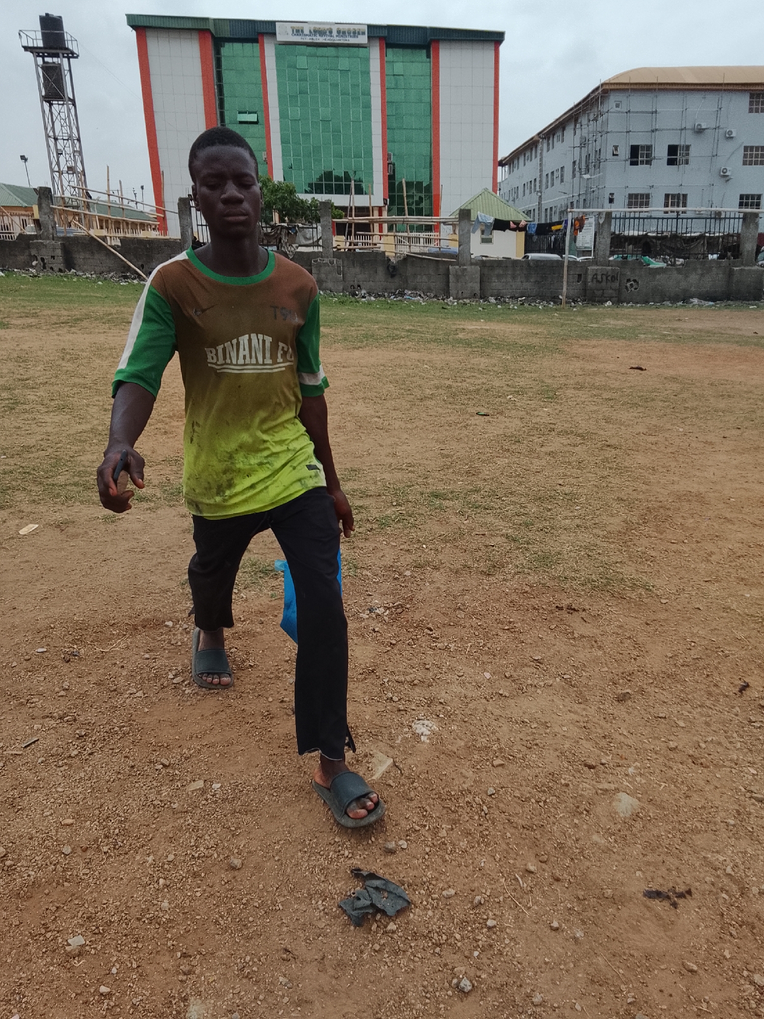 A young man walks across a dirt and grass field. He wears a stained green and brown t-shirt with the word "BINANI" on it, black pants, and open-toed sandals. He carries something in his left hand. Behind him are buildings, including a tall building with green glass and a sign that says "THE LORD'S ORPHANAGE CHARISMATIC REVIVAL MINISTRIES." In the background is a tall metal water tower. The ground is mostly bare dirt with some patches of grass. There is debris on the ground in front of the building and near the man's feet. The photo was taken in the city of Abuja, Nigeria.