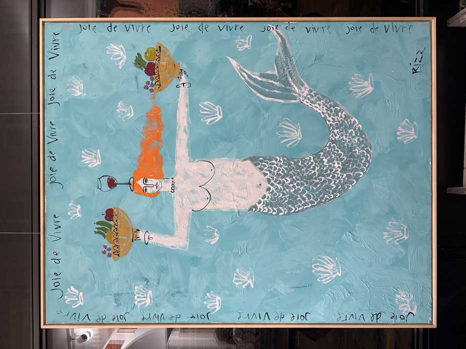 The framed painting features a mermaid with orange hair swimming in a blue sea dotted with white shells. She balances a fruit basket on one hand and a wine glass on her head. The phrase "Joie de Vivre" is repeatedly written along the edges of the painting. The artwork is signed "Rizz" on the upper right corner. The painting's style appears childlike and naive.