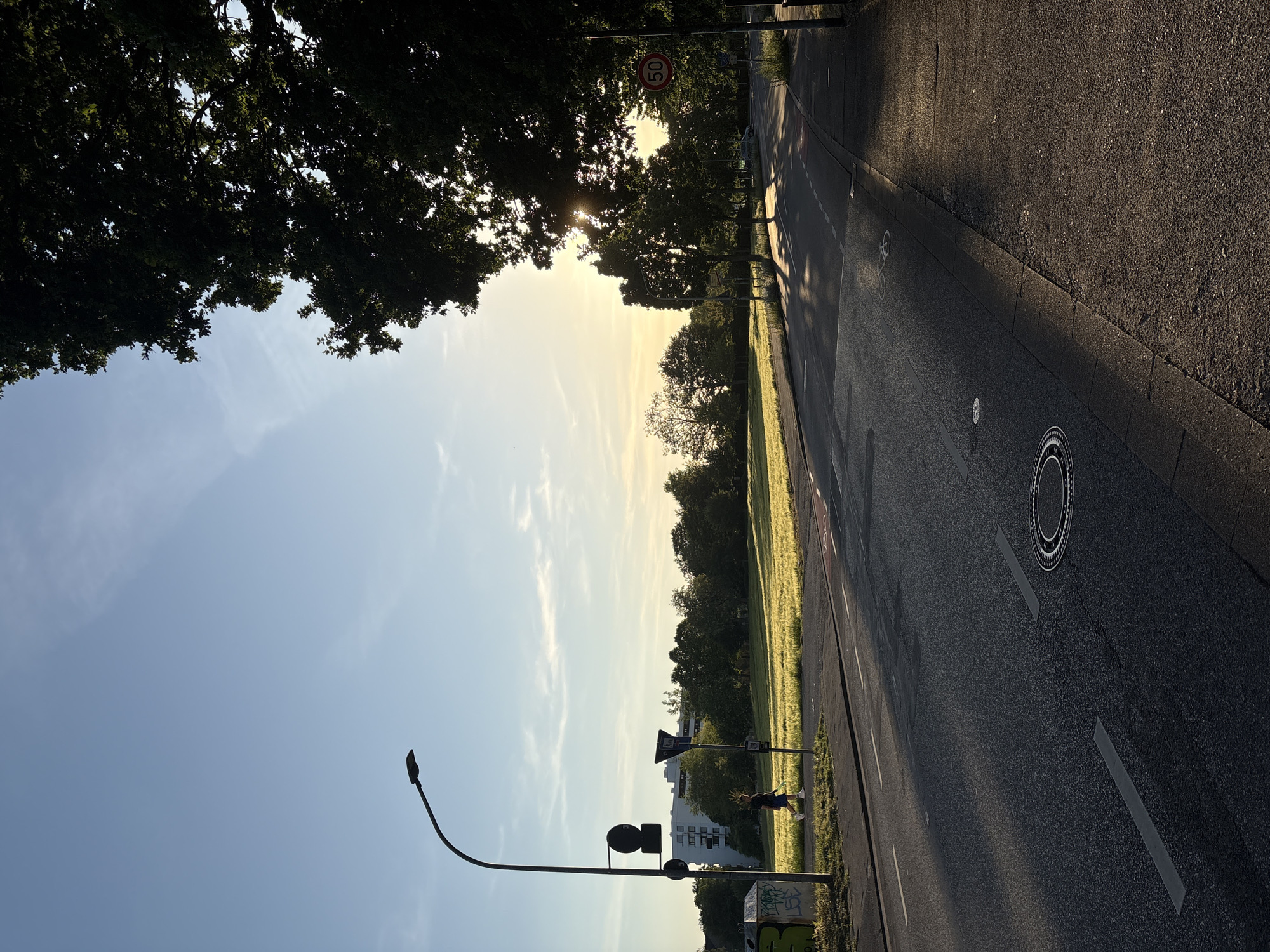 The image shows a street scene in Mönchengladbach, Germany. The road, marked with white lines, runs along a green field or park area. On the left side of the road, there is a lamp post, traffic signs, and some greenery. A person walks past the traffic signs, which are located near buildings. The sky is light blue with some streaks of clouds. Trees border the image on the upper left and further along the street, some obscuring the sunlight. A "50" speed limit sign is visible near the edge of the trees. A manhole cover is visible in the road.