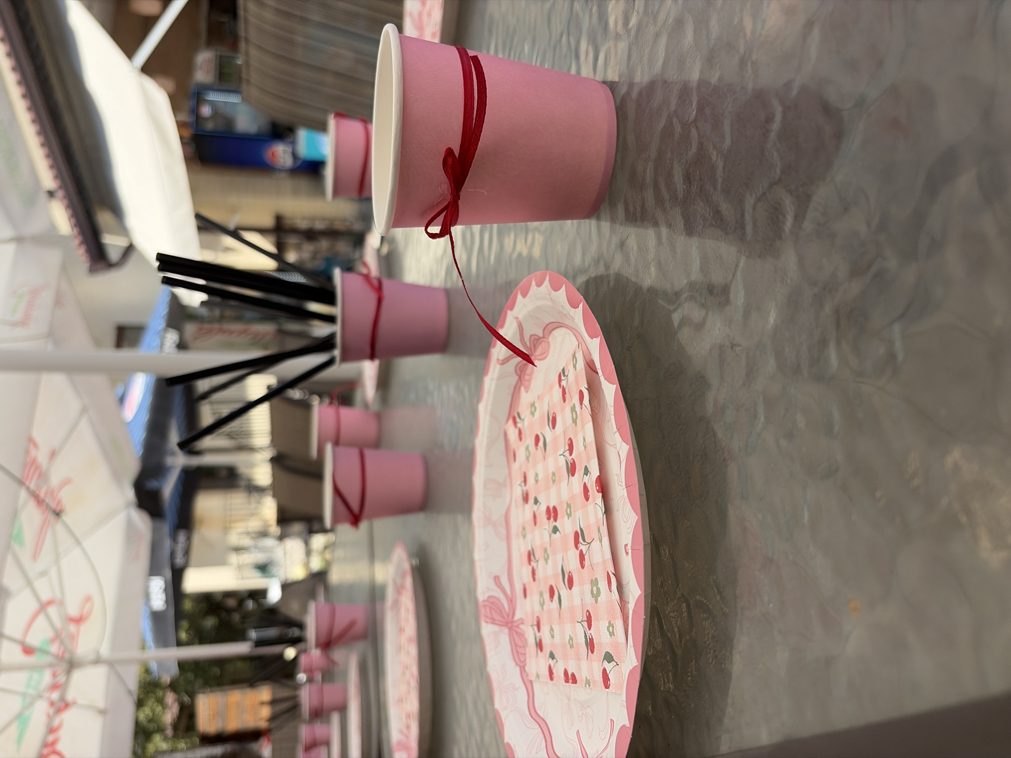 The image shows a table set for an outdoor event, likely a party, in Kolarovo, Bulgaria. The table is covered with a glass-like surface that reflects the surroundings. On the table, there are several pink paper cups, many of which are tied with a thin red ribbon. Some of these cups contain black straws. There are also pink and white paper plates, some of which have a cherry pattern. In the background, there are white umbrellas providing shade, and beyond that, buildings and trees. The scene appears to be set during the daytime, with natural light illuminating the area. There are no people clearly visible in the frame, but the setup suggests a gathering is imminent or has recently concluded. The overall atmosphere is festive and informal, consistent with a celebratory occasion.