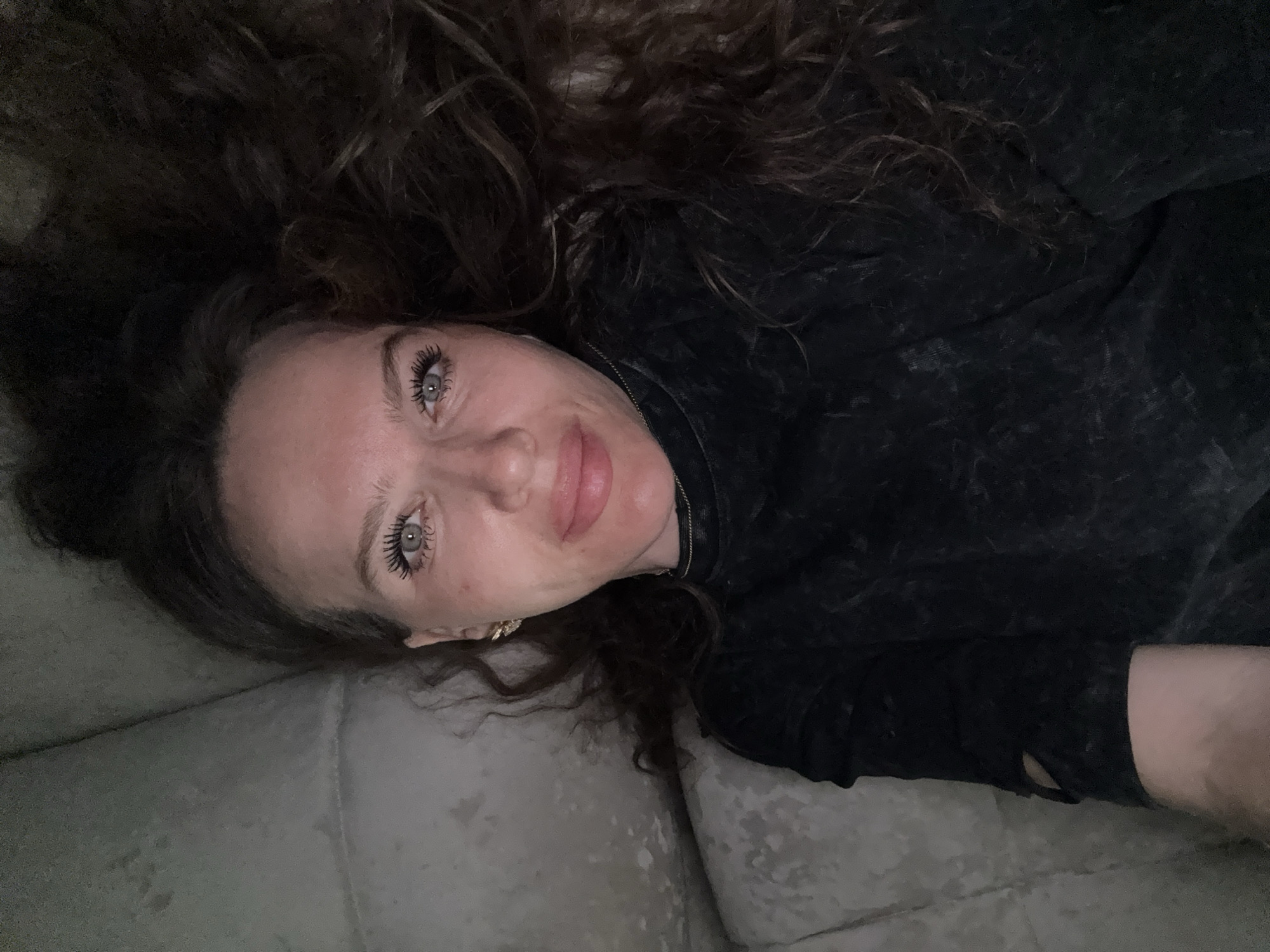 A woman with long, curly brown hair is lying on a gray upholstered surface, likely a couch. She is wearing a dark-colored top, possibly a sweatshirt, and has a visible earring in her left ear. Her eyes are open and she is looking directly at the camera with a slight smile. The location is unknown.