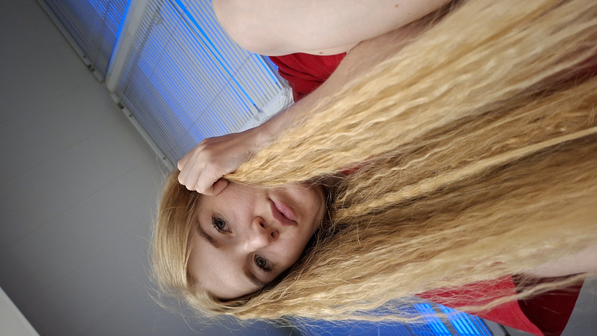 This image is a low-angle, close-up portrait of a young woman with very long, voluminous, wavy blonde hair. She is positioned upside down in the frame, looking directly into the camera with a soft, neutral expression and slightly parted lips. Her eyes appear to be a light color, possibly green or hazel, and she has visible eyelashes. Her right hand is near her head, gently gripping a section of her blonde hair. She is wearing a red garment, likely a top or dress, visible on her shoulder and upper chest.
The background consists of a light gray or white wall/ceiling. A distinctive feature in the upper part of the frame is a set of parallel slats or blinds, illuminated with a bright blue light, suggesting an indoor setting with either a window or a decorative lighting element. The perspective is intimate, taken from below the subject, emphasizing her hair and facial features against the unique blue-lit background.
The activity captured is a posed portrait or selfie. The time of day is indeterminate, but the strong blue light suggests artificial lighting or a specific atmospheric effect within an indoor environment. There is no visible text in the image. The location context provided is Chaikovsky, Russia. The overall mood conveyed is calm and contemplative.