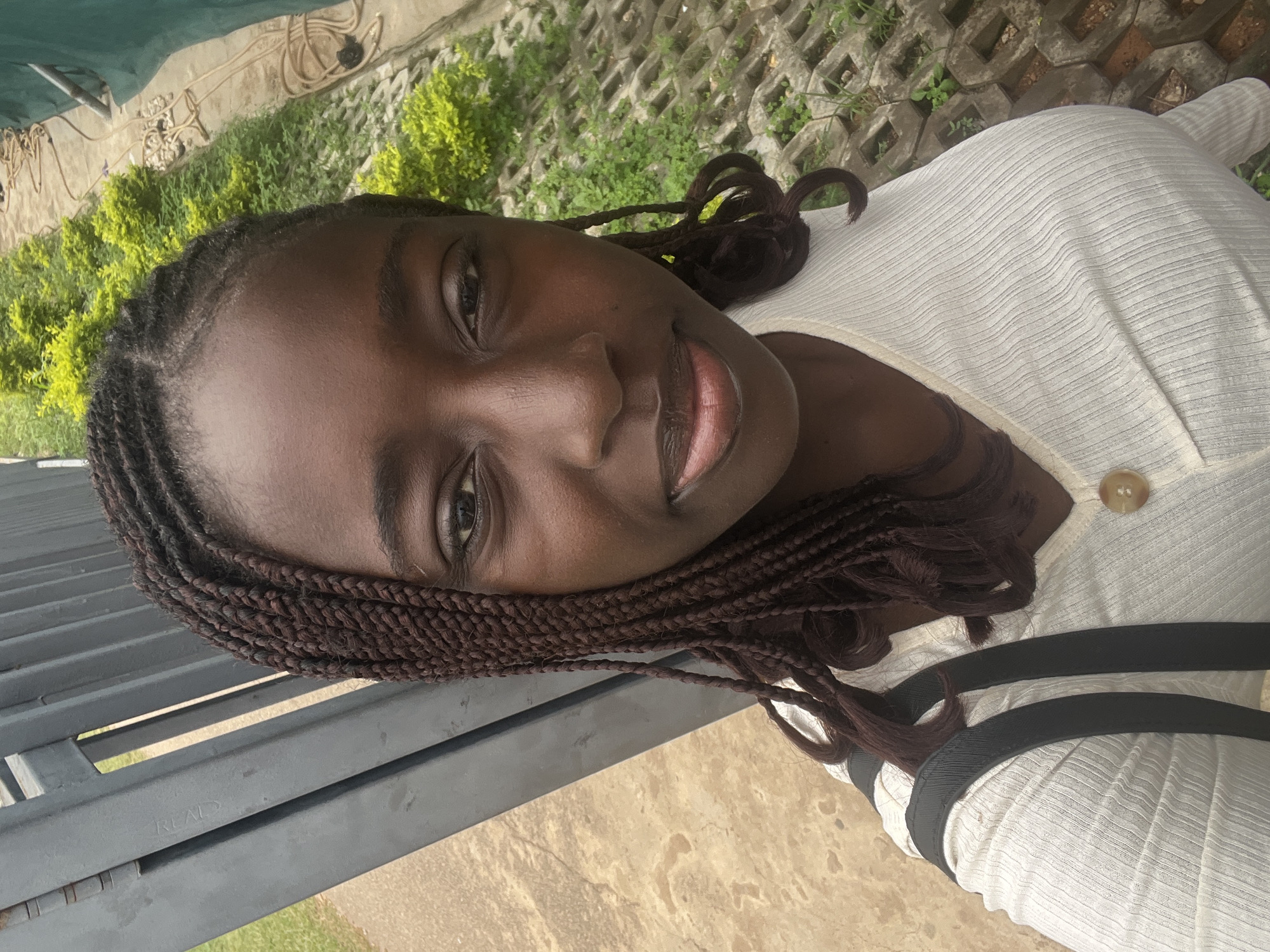 A vibrant, vertically oriented outdoor portrait, taken in Karum, Nigeria, features a young Black woman smiling warmly at the camera. She has a dark complexion and her hair is styled in numerous small, dark brown box braids that frame her face and extend over her shoulders. She is wearing a light-colored, possibly white or cream, ribbed top with a subtle V-neckline and a single light brown button near the collar. A black strap, likely from a bag, is visible across her right shoulder.
The background presents a mix of natural and constructed elements. To her left, a dark grey metal fence or grating is prominently featured, with the word "REAL" faintly visible embossed on one of its beams. Behind her and extending to the right, there's an area with light green, bushy plants and textured concrete blocks or pavers on the ground. Further back, a beige wall is partially obscured by a dark green tarp or awning, with what appears to be coiled pipes or cables affixed to the wall. The lighting is bright and even, indicating a sunny daytime setting, and the overall mood conveyed by the subject's expression is friendly and engaging.