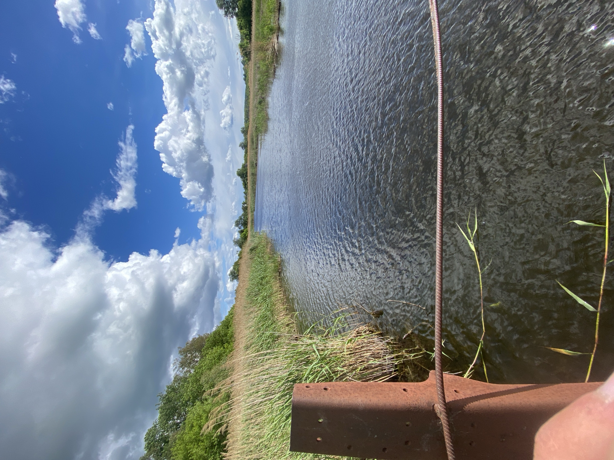The image shows a view of a river or canal from a rusty metal structure, possibly a bridge or dock, in Ustyvytsia, Ukraine. The water is dark and rippled, reflecting the sky. The riverbank is lined with tall grass and reeds, transitioning to trees further inland. The sky is partly cloudy with patches of blue. A metal cable is visible near the edge of the frame. A portion of a finger is visible in the lower right corner.
