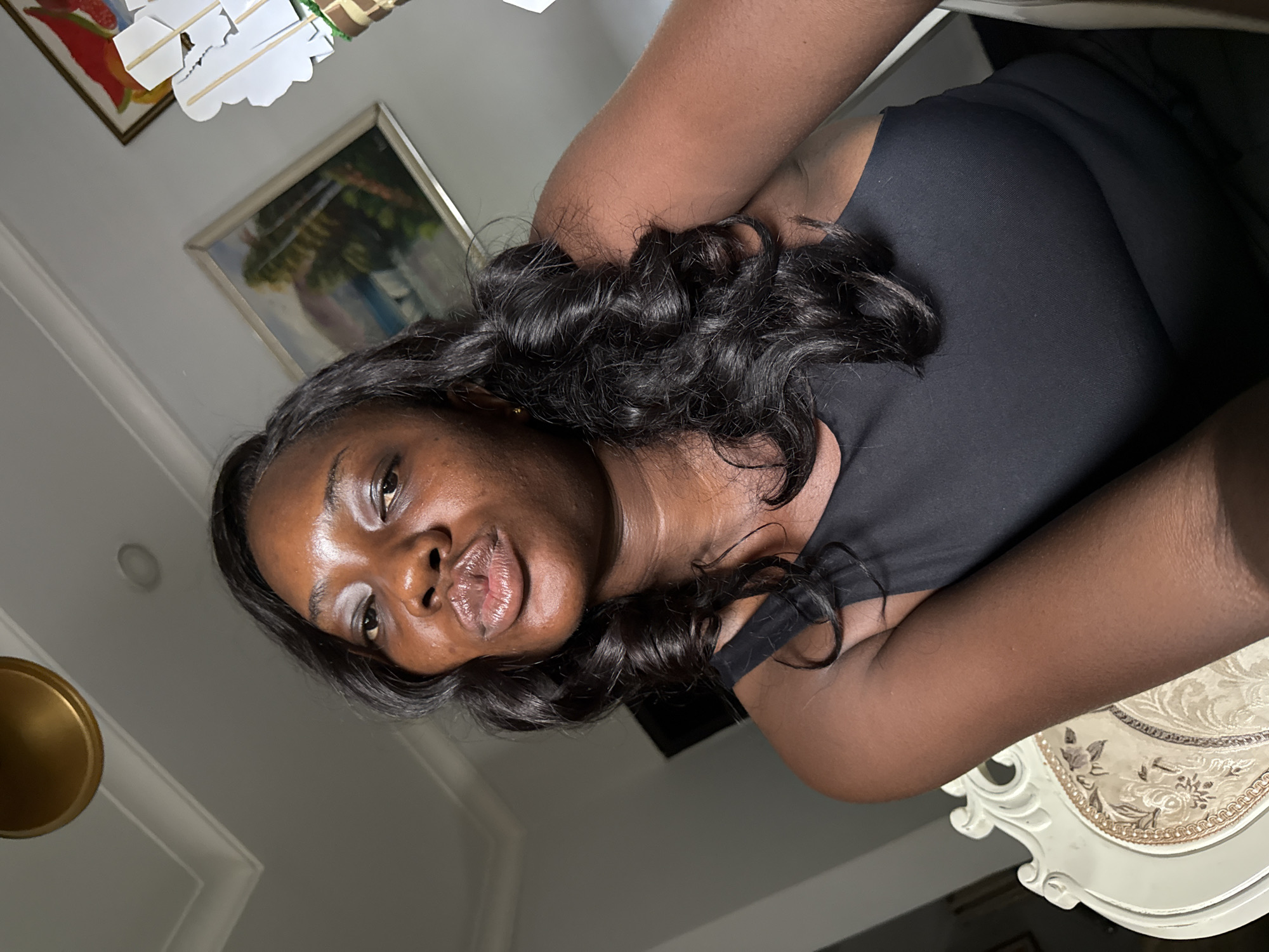 A dark-skinned woman with black, wavy hair is positioned at an angle in the frame. She is wearing a black sleeveless top and appears to be sitting at a white, ornate table. Her arms are crossed. The background includes a framed painting on the wall and a partially visible light fixture. There is no information in the image to determine the location in the city of Iddo Tudunwada, Nigeria.
