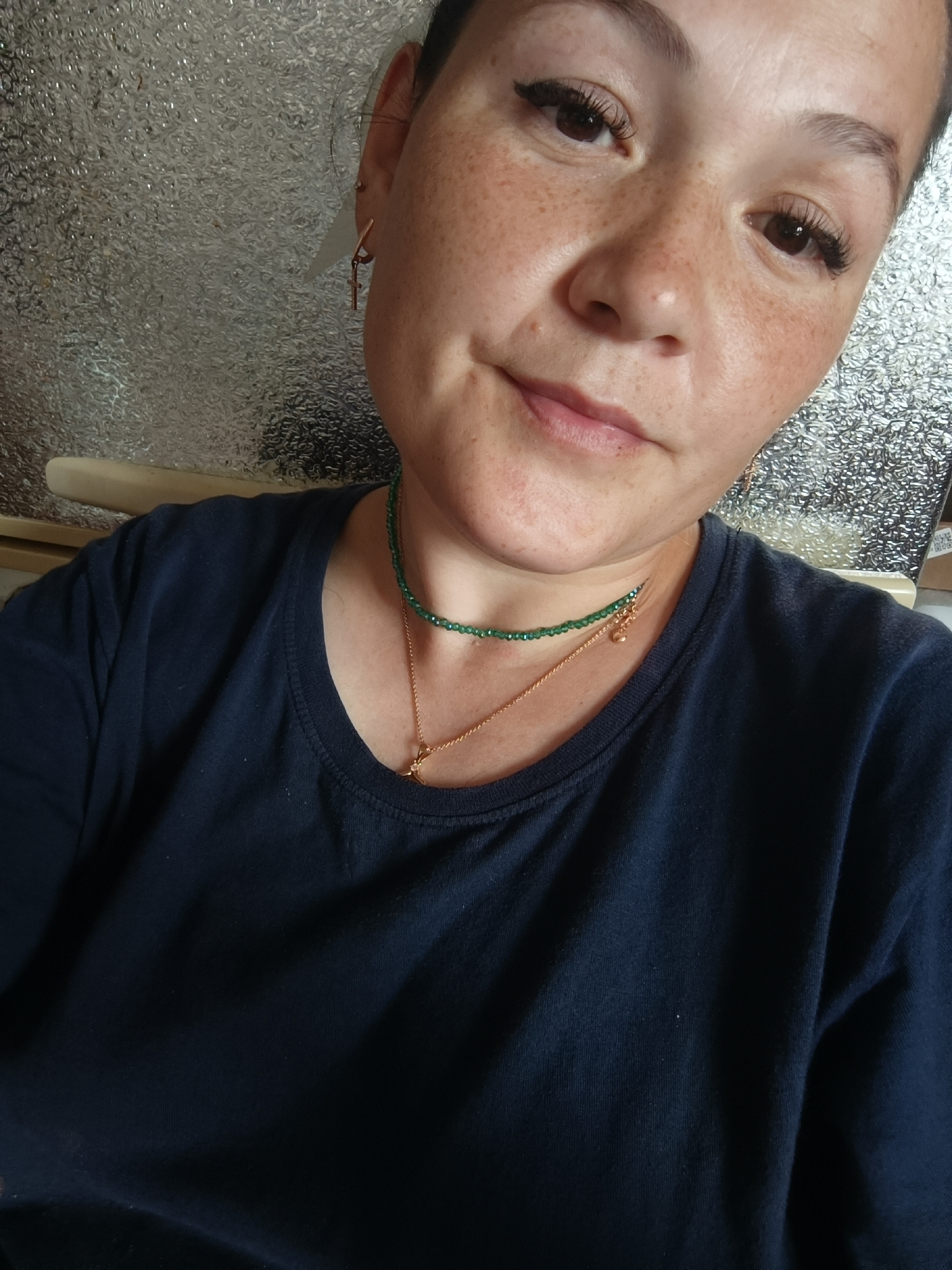The image provided is a close-up selfie of a woman. She has fair skin with freckles, dark hair pulled back, and is wearing makeup, including winged eyeliner and false eyelashes. She is adorned with two necklaces, one a green beaded choker and the other a gold chain with a pendant. She is wearing a dark blue t-shirt. The background appears to be a textured, reflective surface. There is no information within the image that allows for the determination of its location in the city of Karsha, Russia.