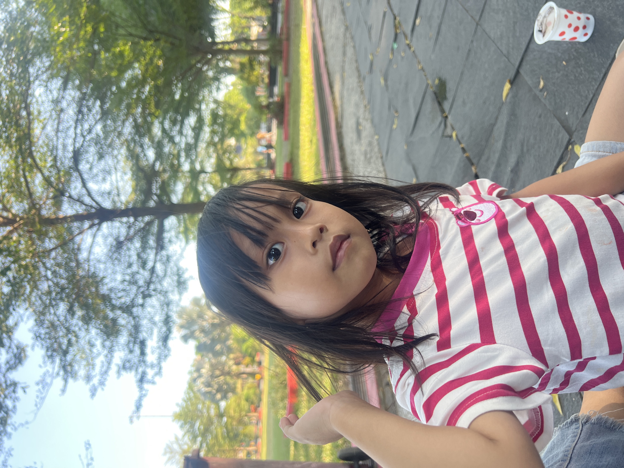 The image is a close-up, low-angle shot of a young girl outdoors. She has dark hair and is looking directly at the camera with a neutral expression. She is wearing a white t-shirt with bold pink stripes and what appear to be denim shorts. Her left hand is extended towards the viewer.
In the background, trees with dappled sunlight filtering through their leaves create a soft, out-of-focus effect. The ground appears to be paved with dark tiles. To the right of the girl, a small, white cup with red polka dots is visible on the ground. The scene suggests a park or playground setting, possibly on a sunny day. The lighting indicates it is likely daytime. The overall mood is relaxed and candid. No visible text is present in the image. The location context is Purwodadi, Indonesia.
