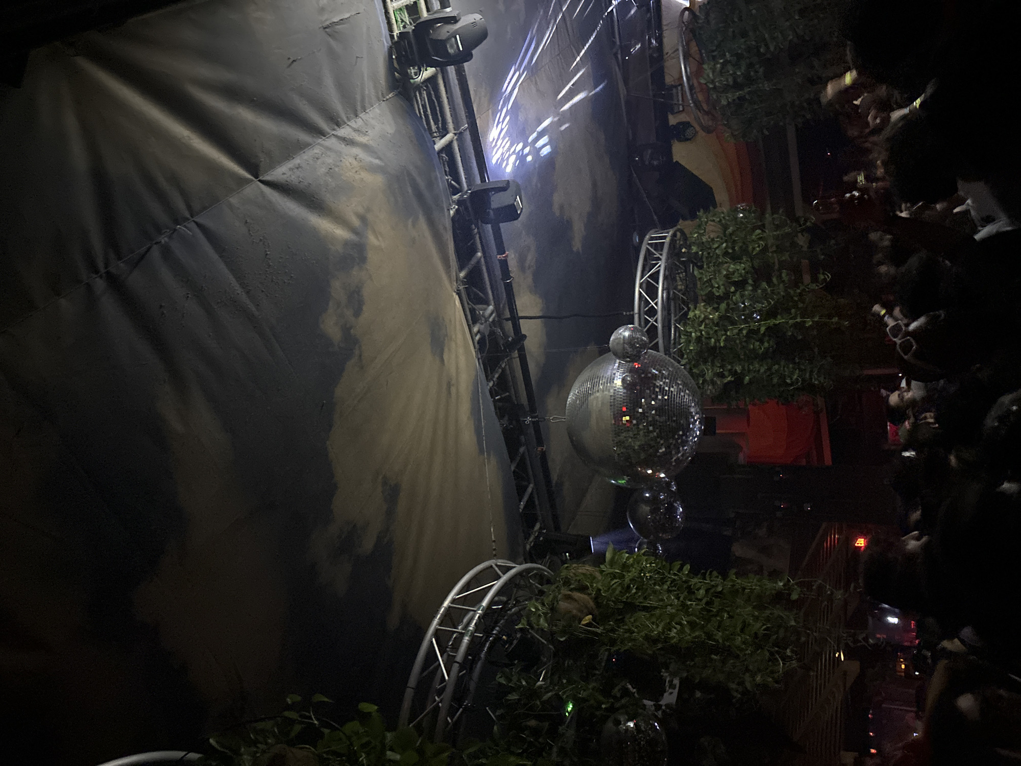 The image captures the interior of a crowded entertainment venue, identified as The Dirty Rabbit Wynwood in Miami, United States. The upper half of the frame features a dark, textured fabric ceiling patterned with lighter, cloud-like forms, displaying visible creases and seams. Suspended from this ceiling are multiple silver metal truss structures. Several stage lighting fixtures, including moving heads, are mounted on these trusses, with one actively projecting bright, patterned beams onto the ceiling surface. Three disco balls of different sizes hang centrally, reflecting ambient light. Lush green hanging plants, likely artificial, are draped extensively over the truss structures and around the disco balls, adding to the decor. The lower half of the image is densely populated by a crowd of people, mostly visible as dark silhouettes from the shoulders up, suggesting a lively social gathering. A blurred red "EXIT" sign is dimly visible in the background towards the left. The overall scene is dimly lit, focusing on the illuminated decorations and the large gathering of individuals.