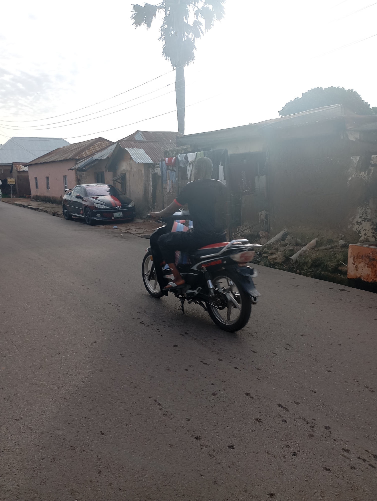 A person rides a motorcycle down a street in Shendam, Nigeria. The person is wearing a black t-shirt and black pants, and appears to be male. The motorcycle is red and black, with silver accents. The street is paved with asphalt, and there are houses on either side of the street. There is a car parked on the side of the street. The sky is cloudy. The environment is an urban setting.